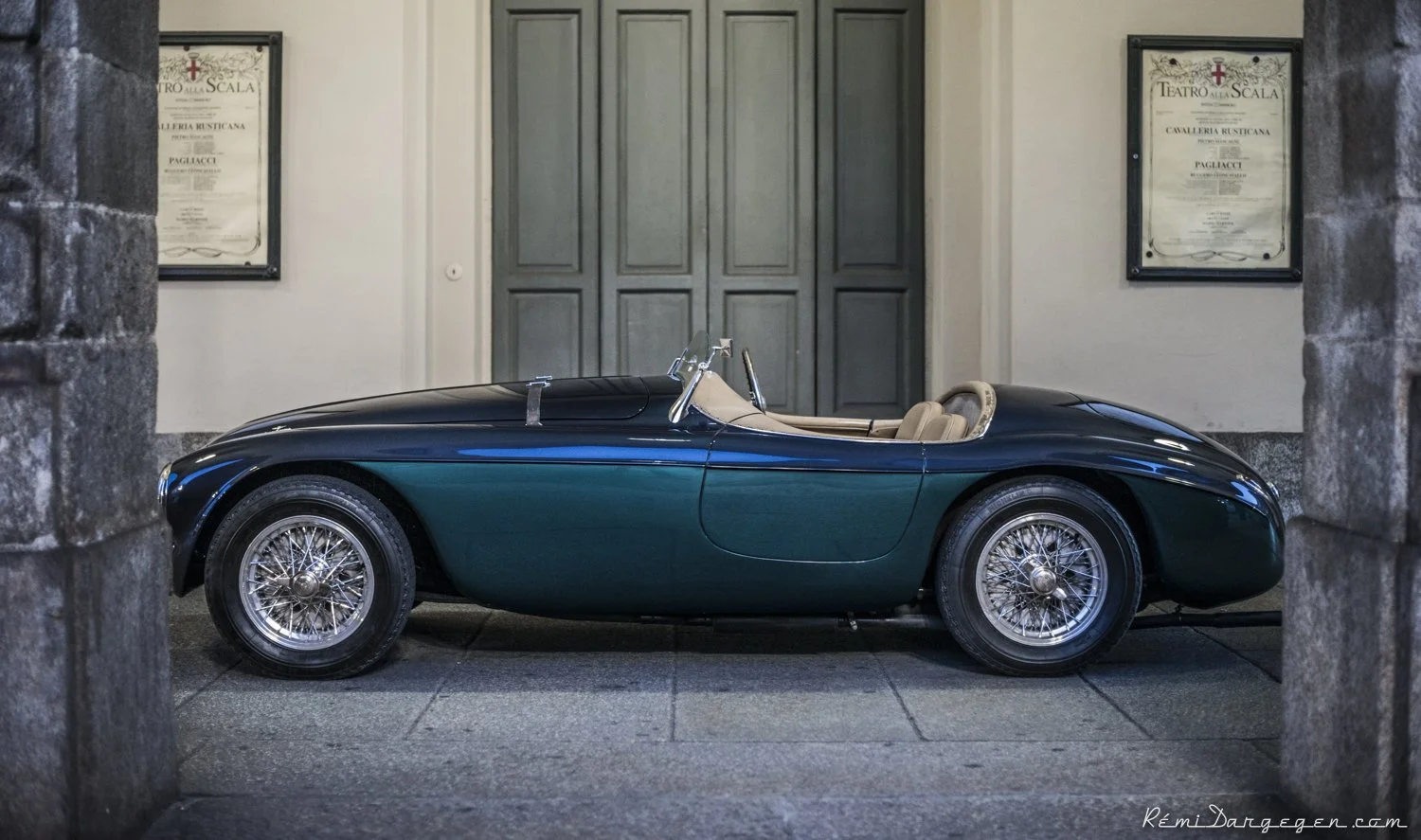 A vintage convertible sports car with a dark green body and beige interior parked indoors, framed by stone columns and stone floors, with framed posters on the wall in the background.
