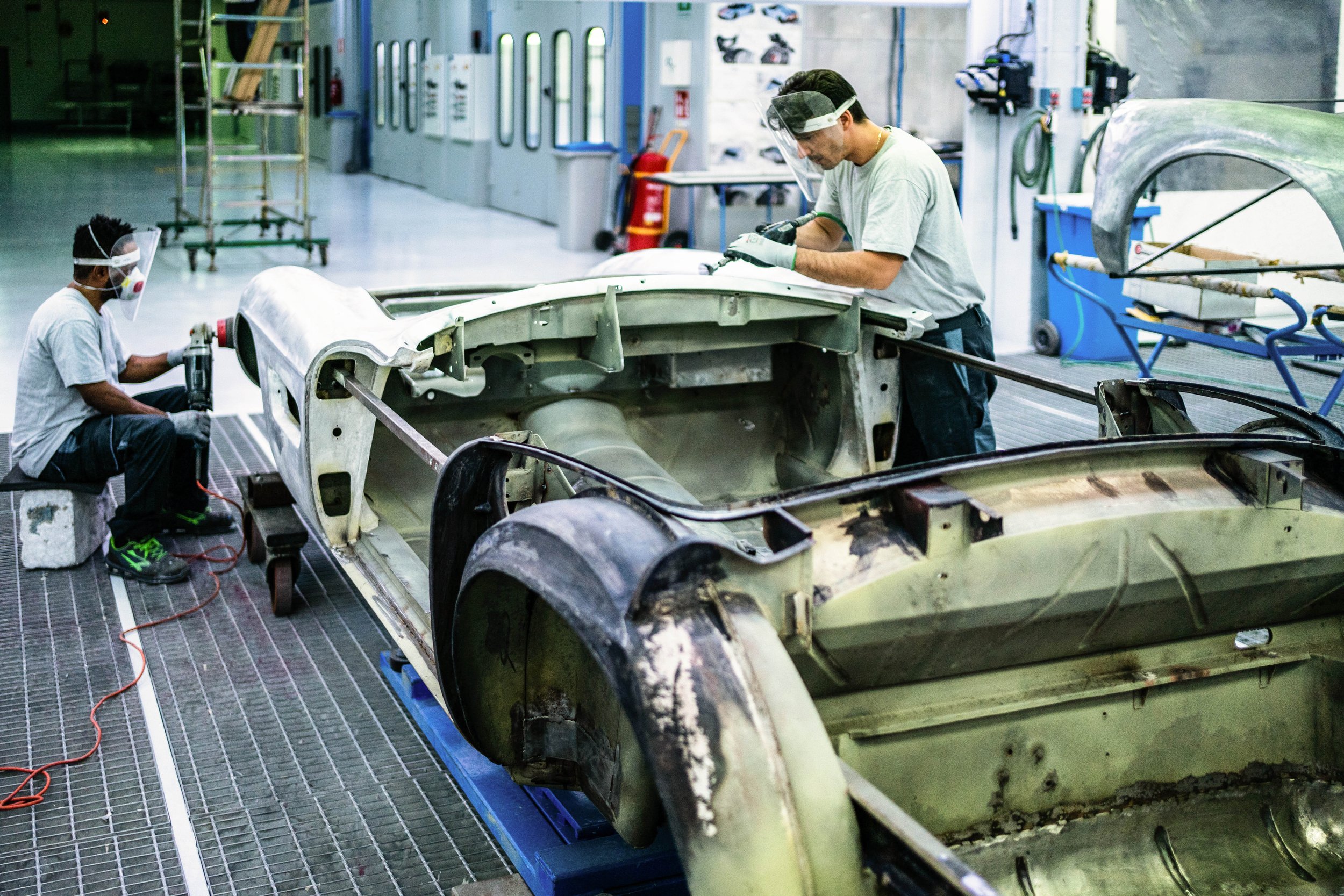 Two workers are repairing a vintage car in an auto restoration shop. One is using a power tool on the car's body near the front wheel, while the other is examining or working on the interior of the car.