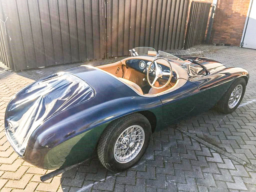 A vintage black race car with a tan interior parked on a brick driveway, next to a brown wooden fence.