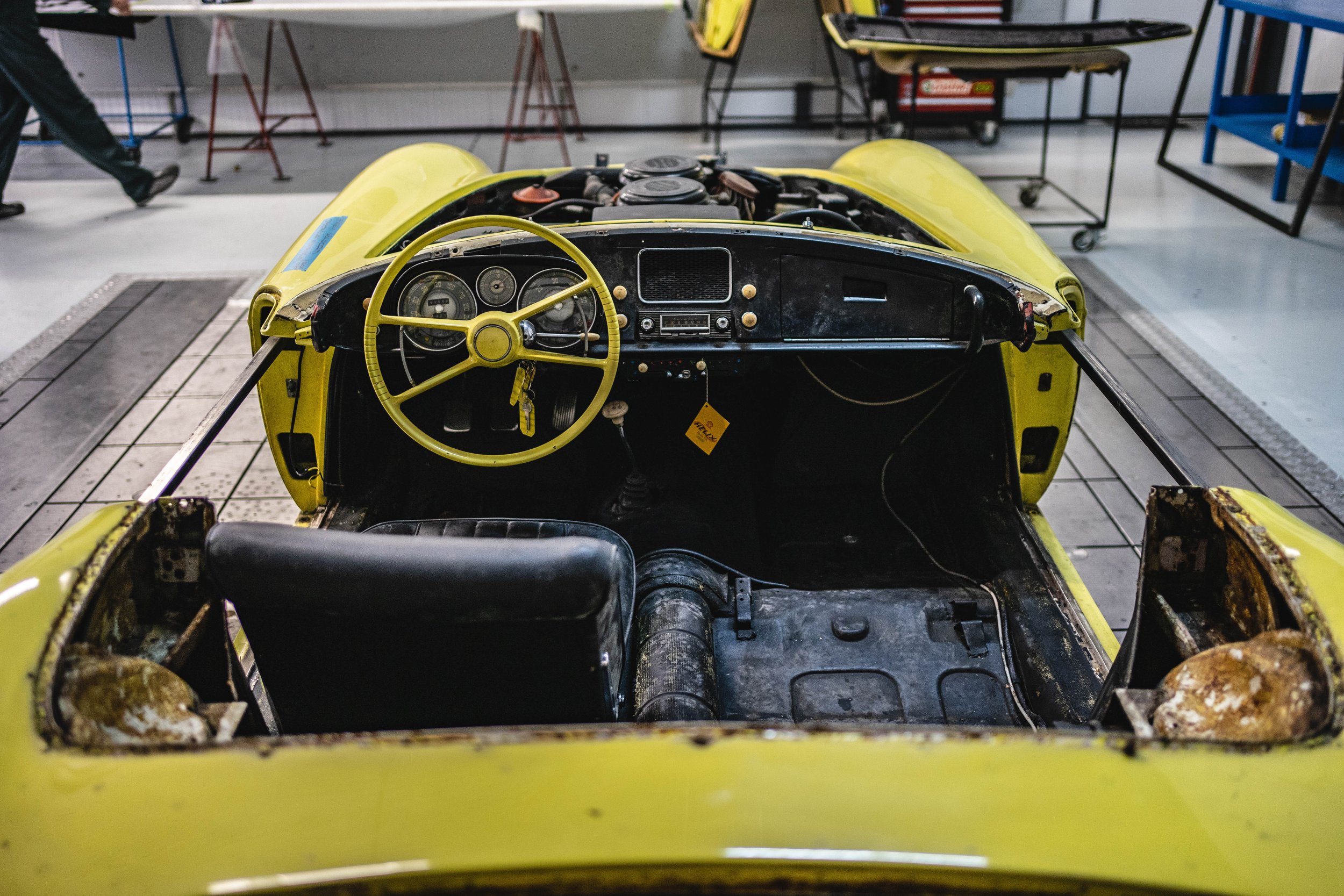 View of an old yellow race car or sports car interior in a workshop, showing a yellow steering wheel, black dashboard, and worn seats with visible rust and damage.