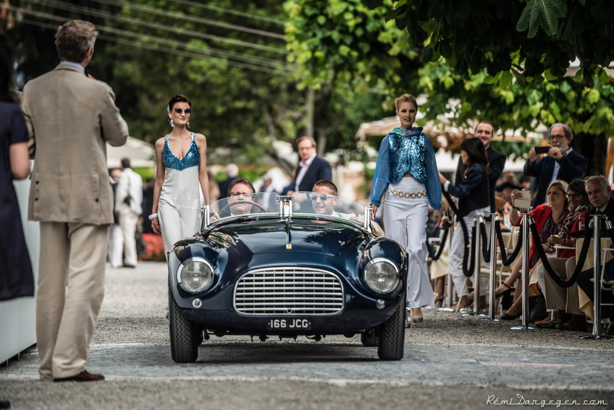 A vintage black convertible car with a license plate reading 166 JCG driving through a crowded outdoor event with people seated on both sides. The scene is set under green leafy trees, with some attendees taking photos and wearing stylish outfits.