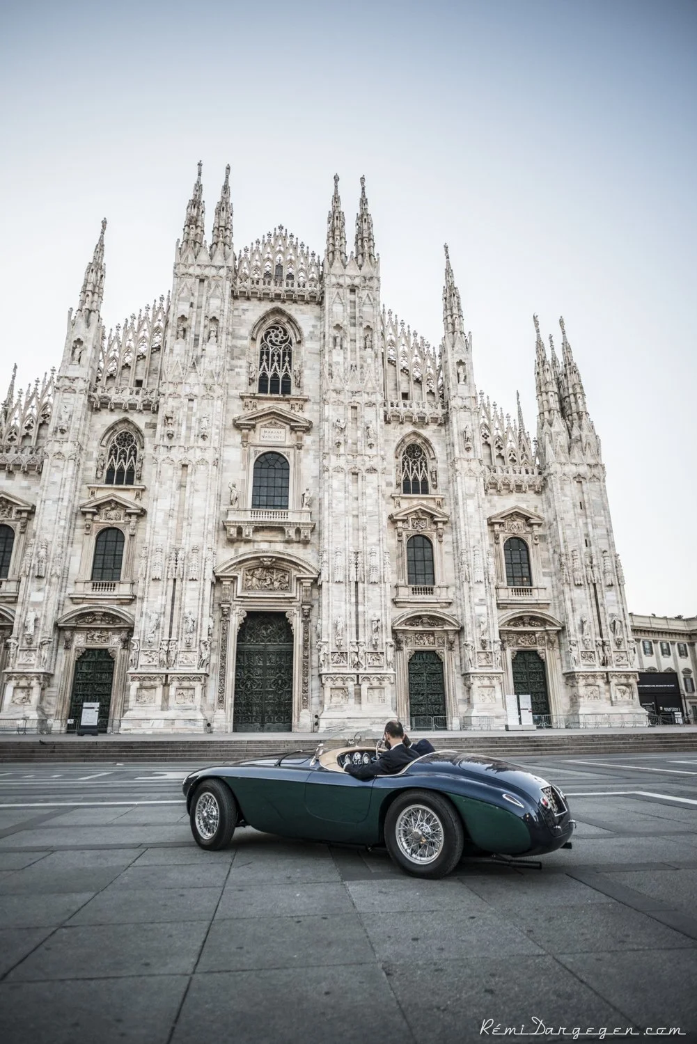 A vintage green and black convertible car parked on a city street in front of the Milan Cathedral in Italy, with a man in the driver’s seat.