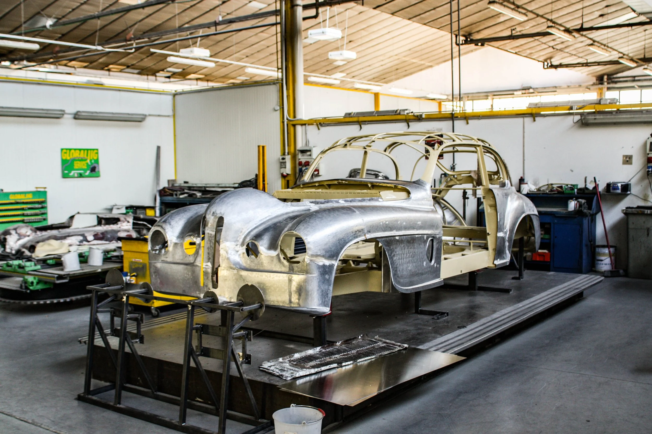 A vintage race car under restoration in a workshop, with its body panels and framework visible, surrounded by various tools and equipment.
