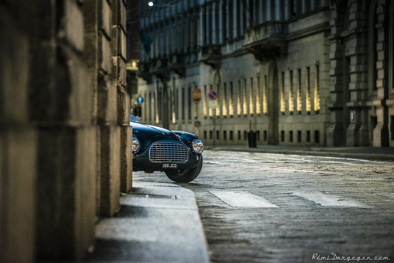A vintage black car is parked on a cobblestone street, partially visible behind a stone wall, in an urban setting with old buildings and a no parking sign in the background at dusk or early evening.