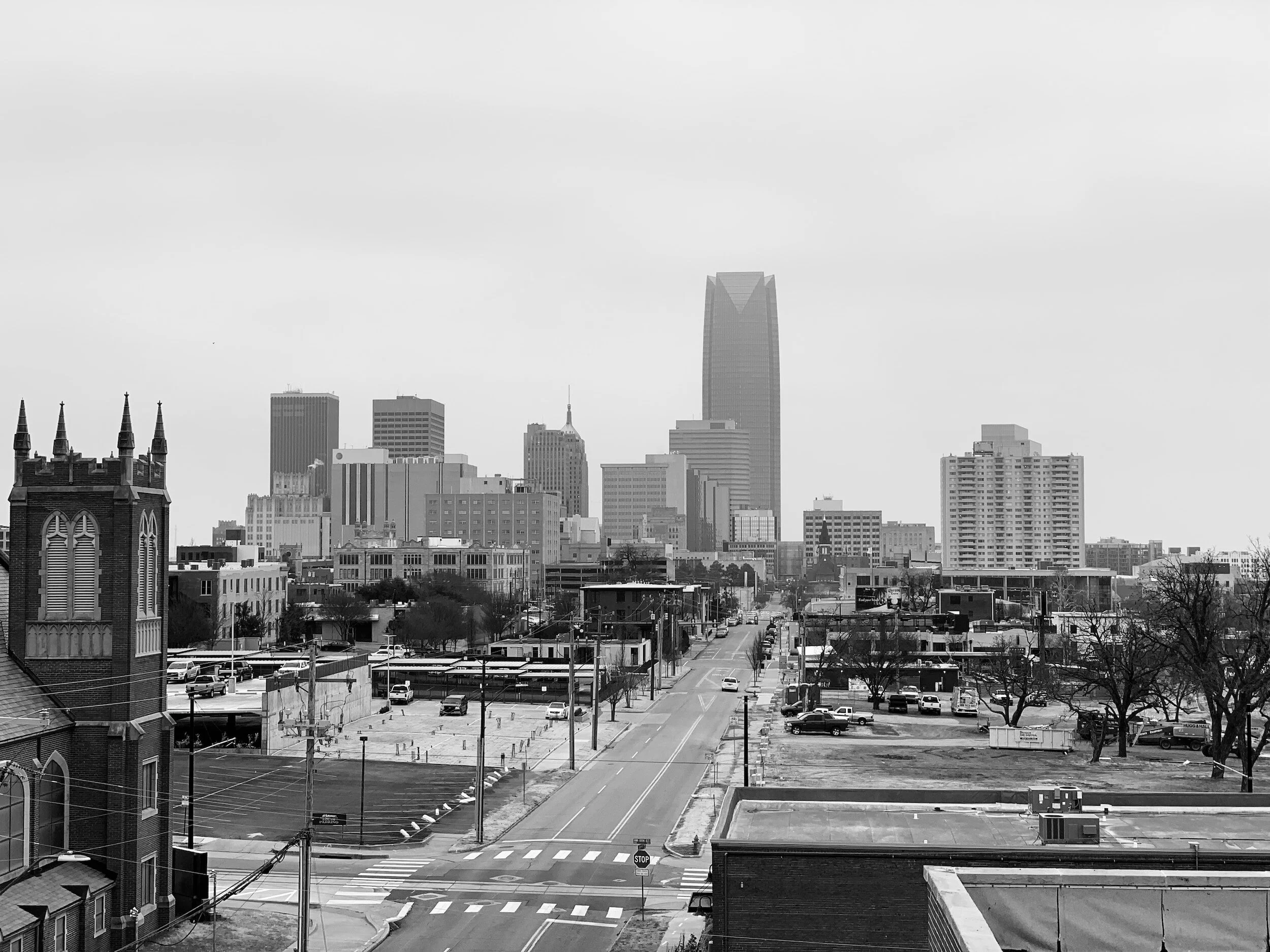 Black and white photo of downtown cityscape with various tall buildings, street, parked cars, trees, and a church with steeples in the foreground.
