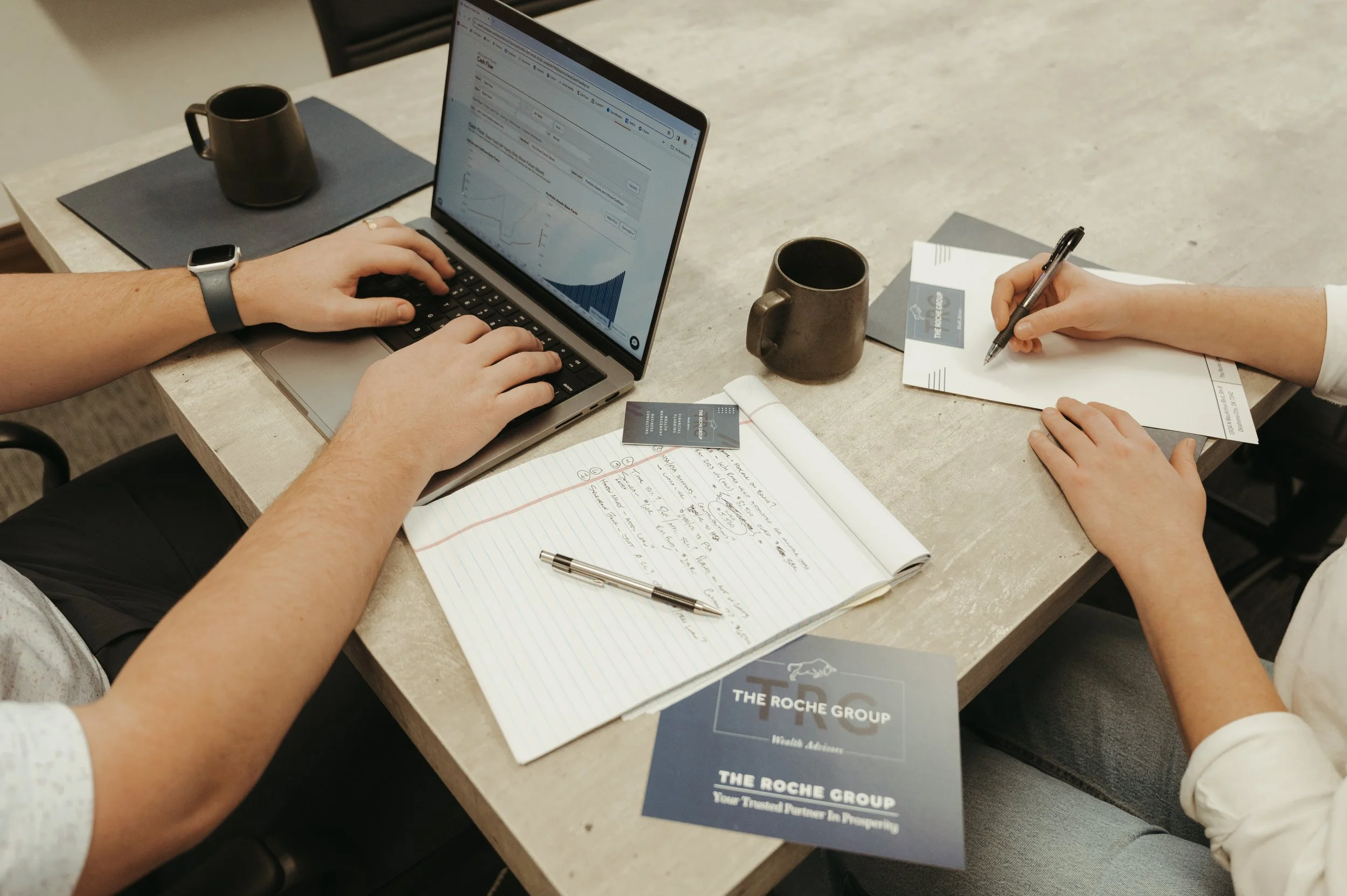Two people working at a table with a laptop, notebooks, and coffee mugs, with business cards from 'The Roche Group'.