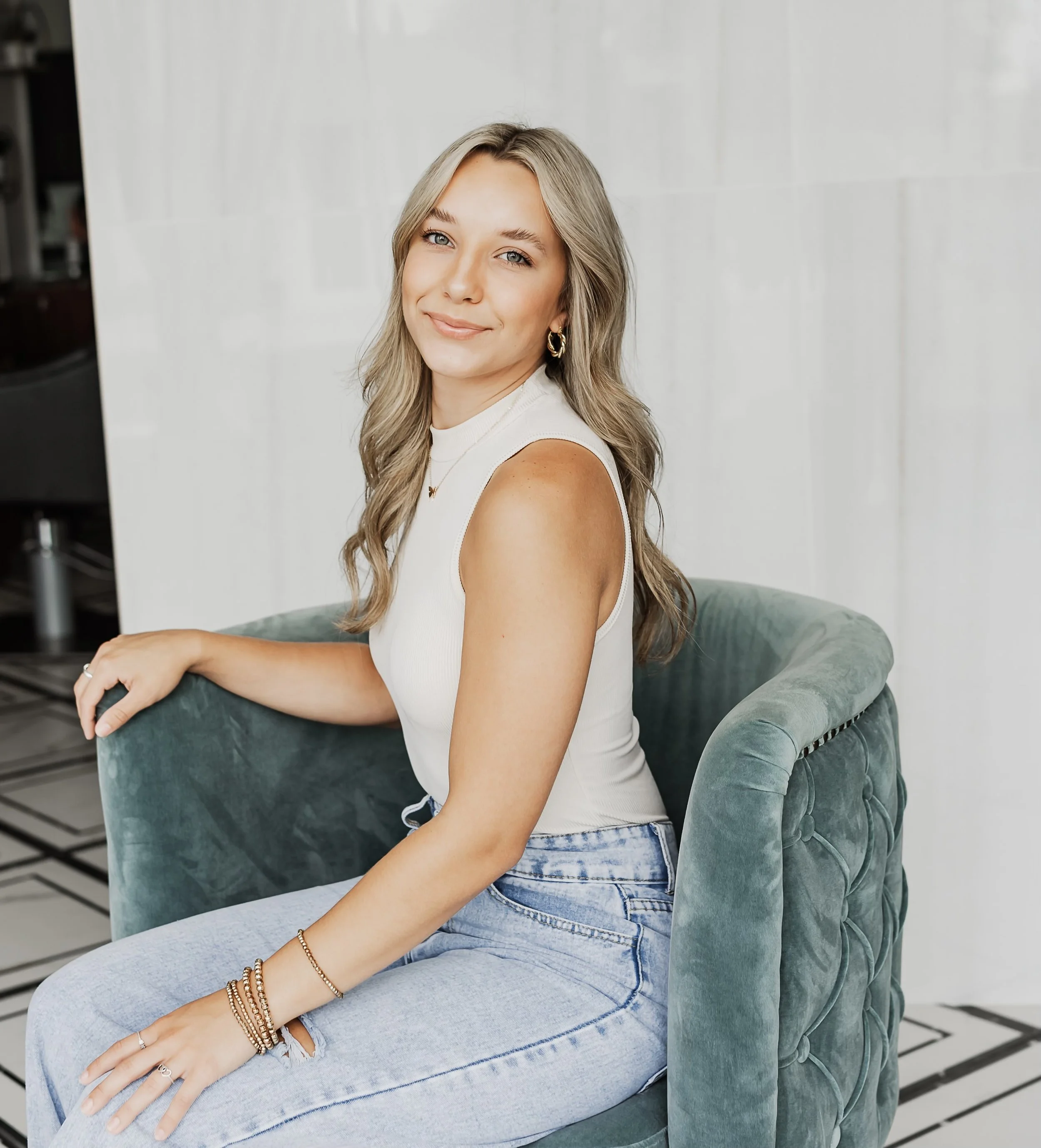 A smiling woman with long blonde hair sitting sideways on a green upholstered armchair, wearing a white sleeveless top, light blue jeans, and jewelry, in a modern indoor setting.