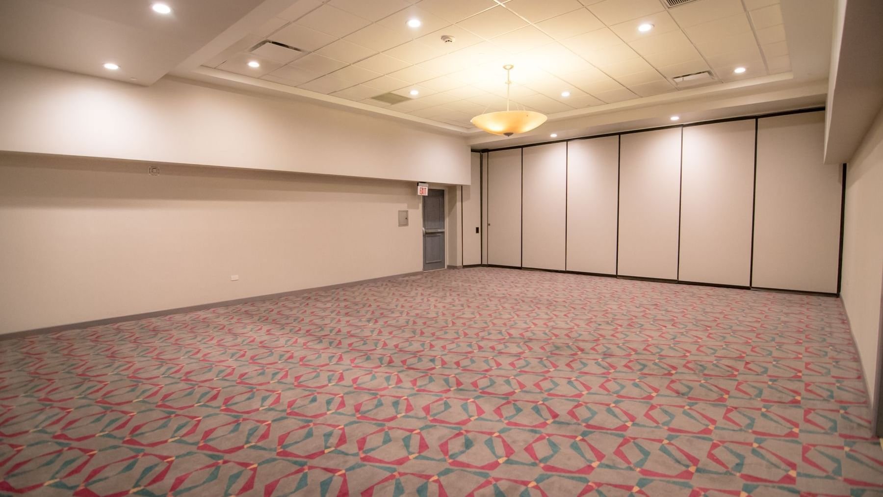 Empty banquet or conference room with patterned carpet, plain walls, a ceiling light, and a door near the corner.