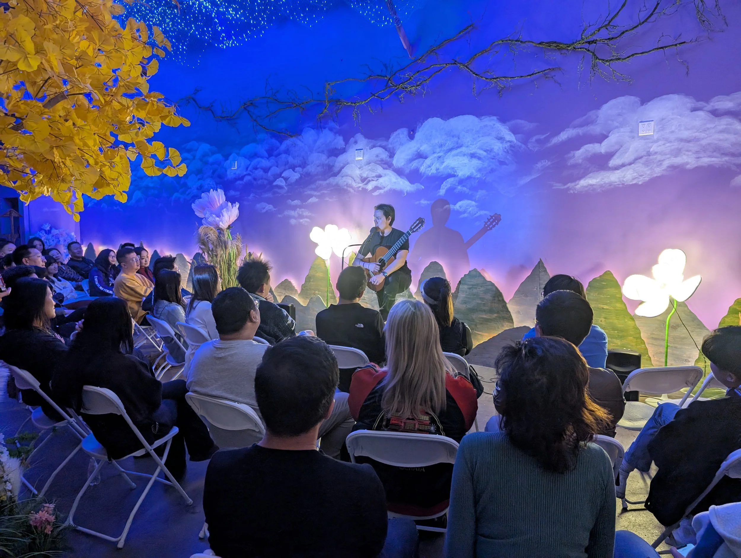 A live music performance with a singer playing guitar on stage, surrounded by decorative flowers and scenic backdrop with clouds and mountainous terrain, audience seated and watching.