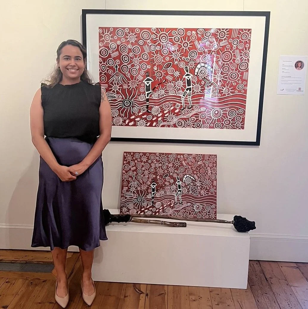 A woman standing in front of two paintings with abstract red and white circular designs, and a black and brown staff or cane resting on a white display platform.