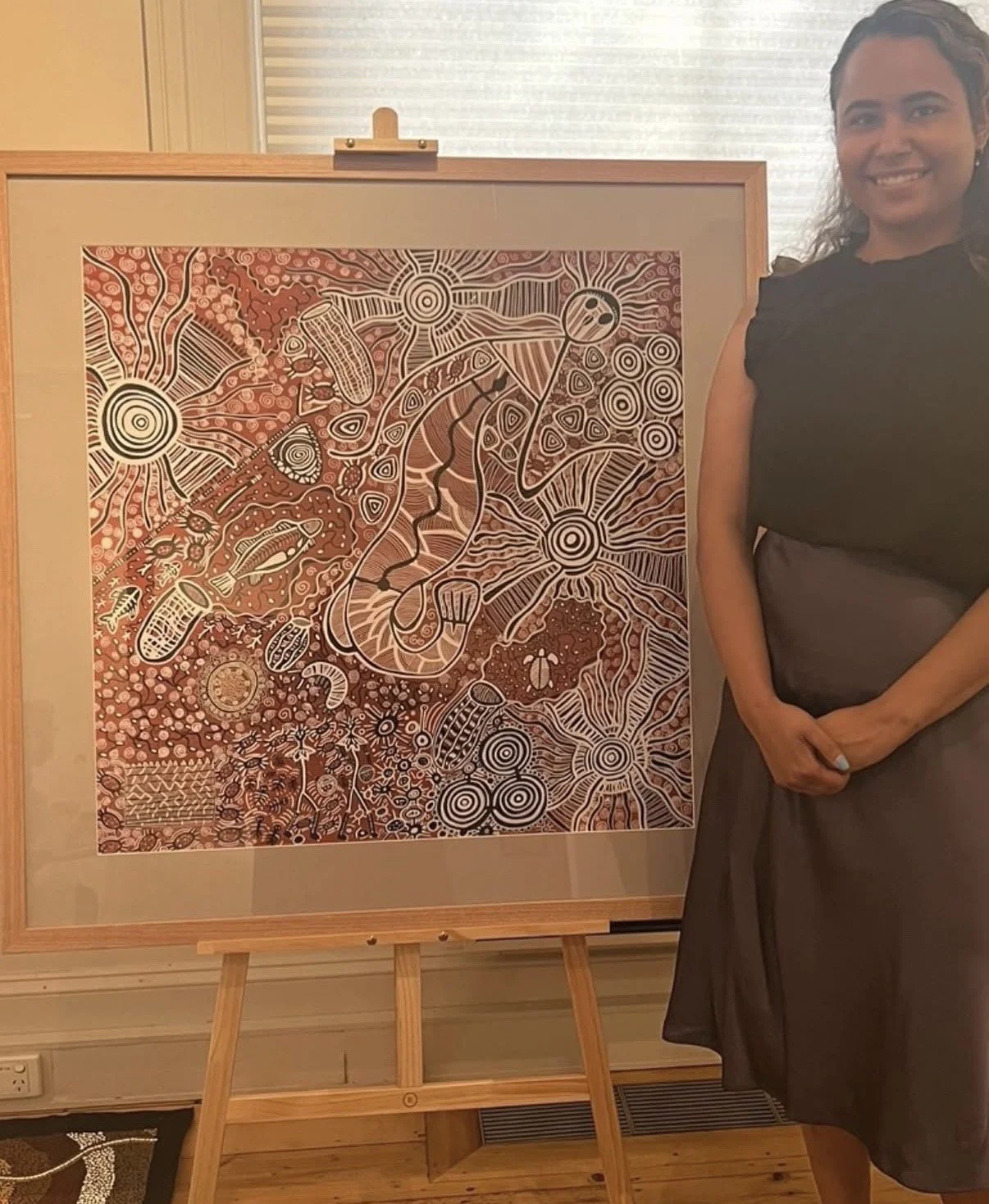 A woman in a black top and gray skirt stands next to an artwork on an easel. The artwork features intricate patterns and designs in black, white, and shades of red or brown.