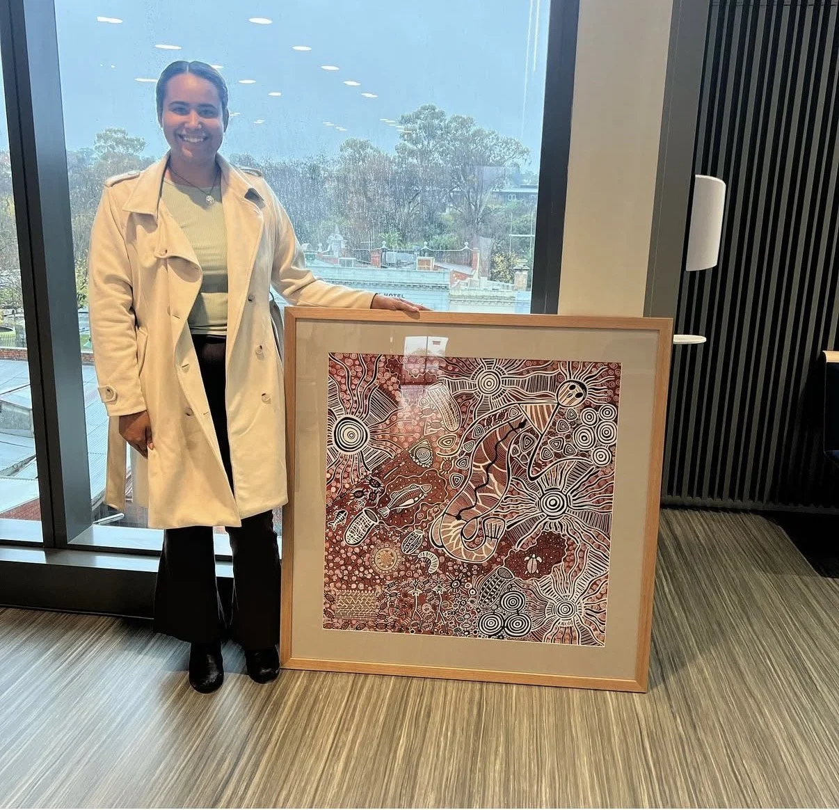 A woman with dark hair wearing a beige coat and black pants standing indoors beside a framed colorful artwork with intricate patterns and animal figures, near a large window showing trees and buildings outside.