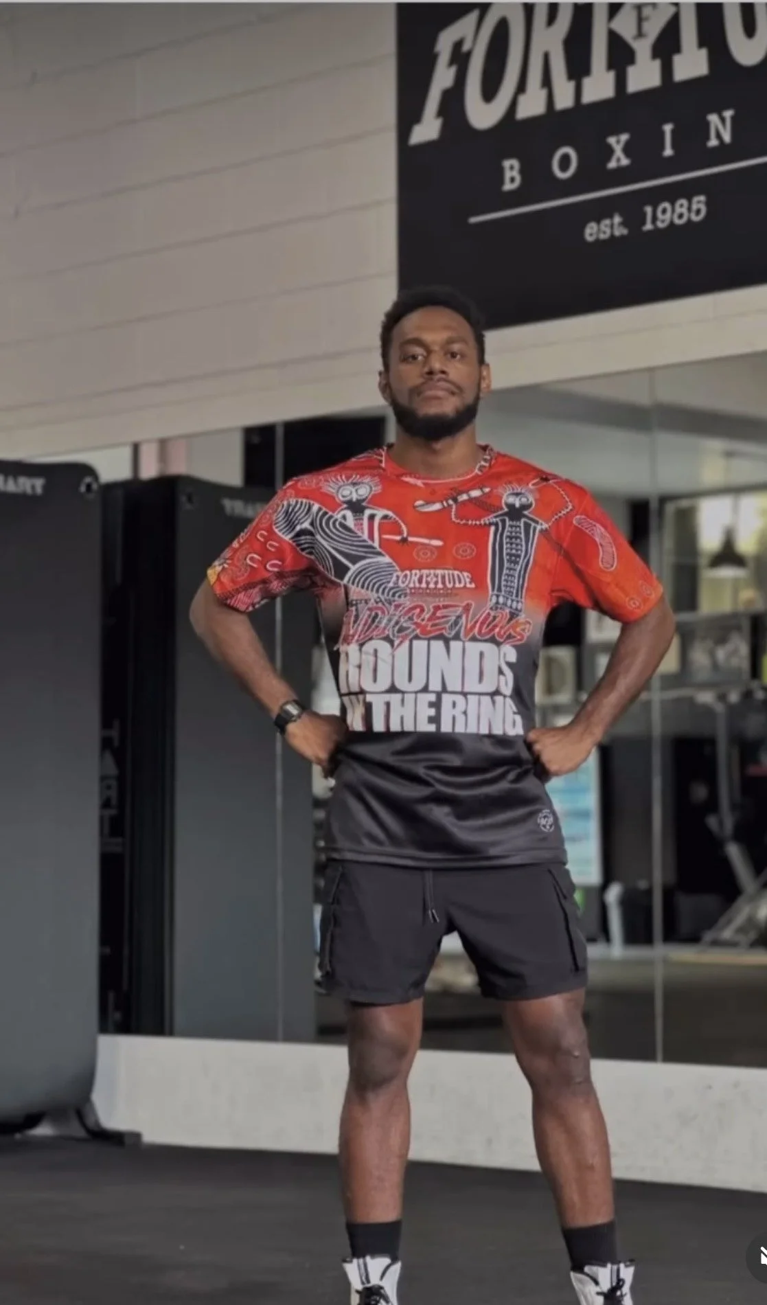 A man standing confidently in a gym, wearing a colorful sports shirt and black shorts, with hands on hips, in front of boxing equipment and a mirror with a sign that reads 'Fortitude Boxing.'