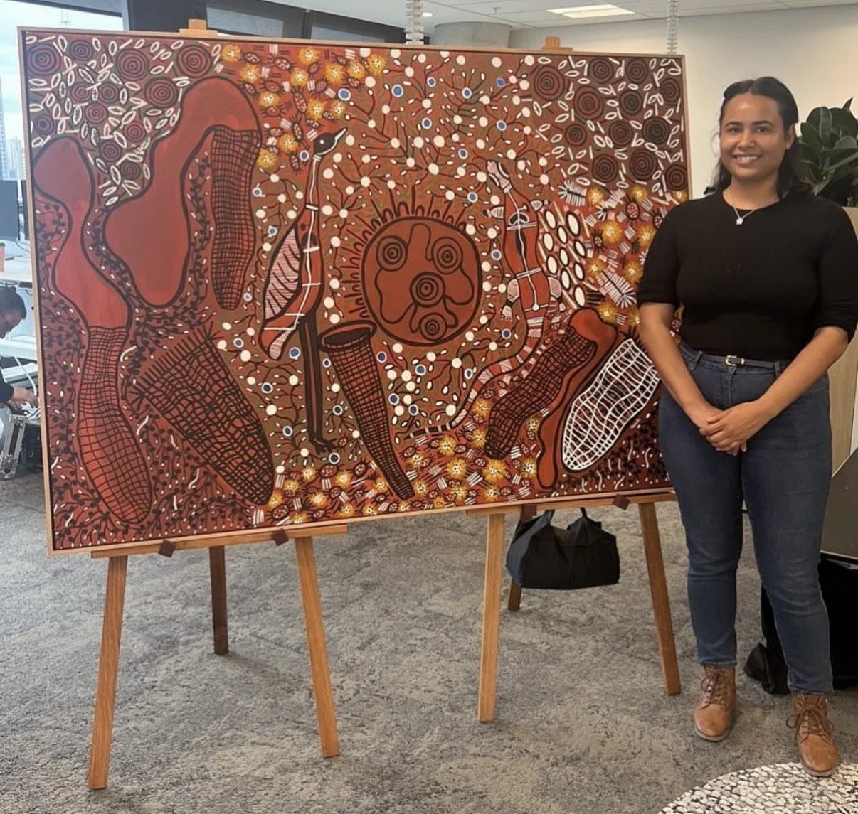 A woman standing next to a colorful Aboriginal-style painting on an easel. The painting features traditional patterns, dots, and abstract figures in red, brown, black, white, and yellow tones.