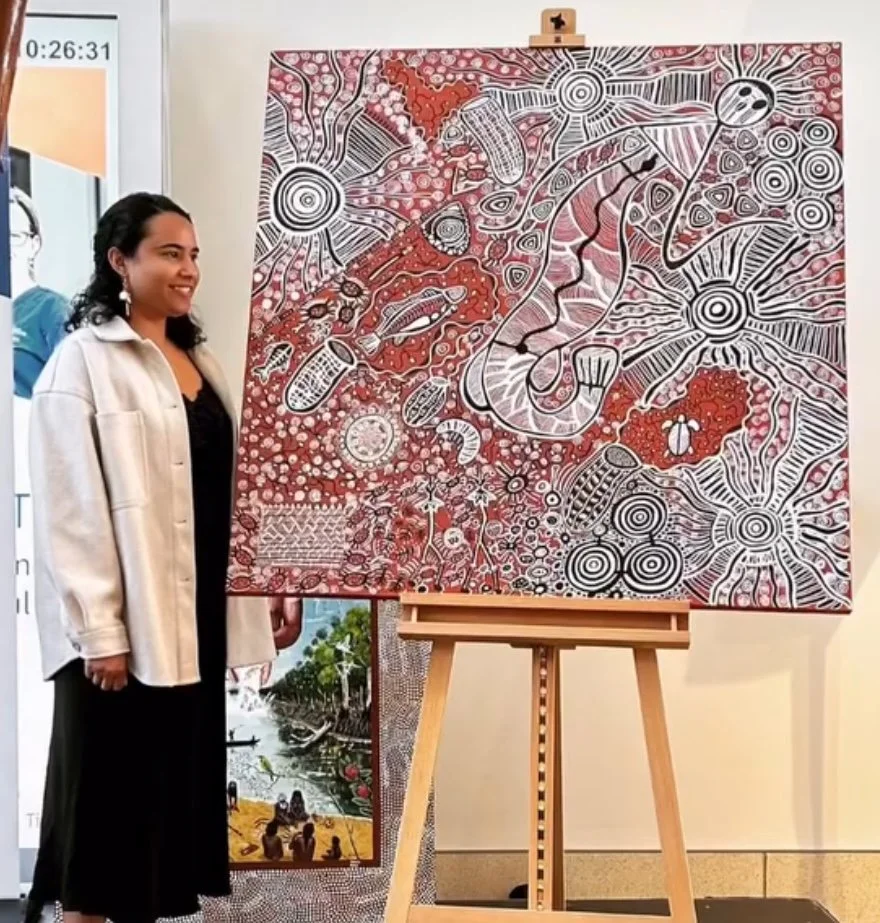 A woman standing next to a large abstract artwork on an easel. The artwork features red, black, and white colors with intricate patterns, circles, and shapes. There is a smaller landscape painting on the floor beside her.