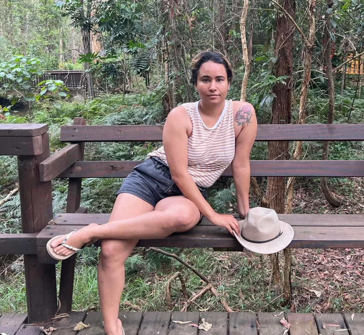 A young woman with dark hair and a tattoo on her shoulder sitting on a wooden bench in a forest, holding a beige hat, wearing a striped tank top, shorts, and flip-flops.