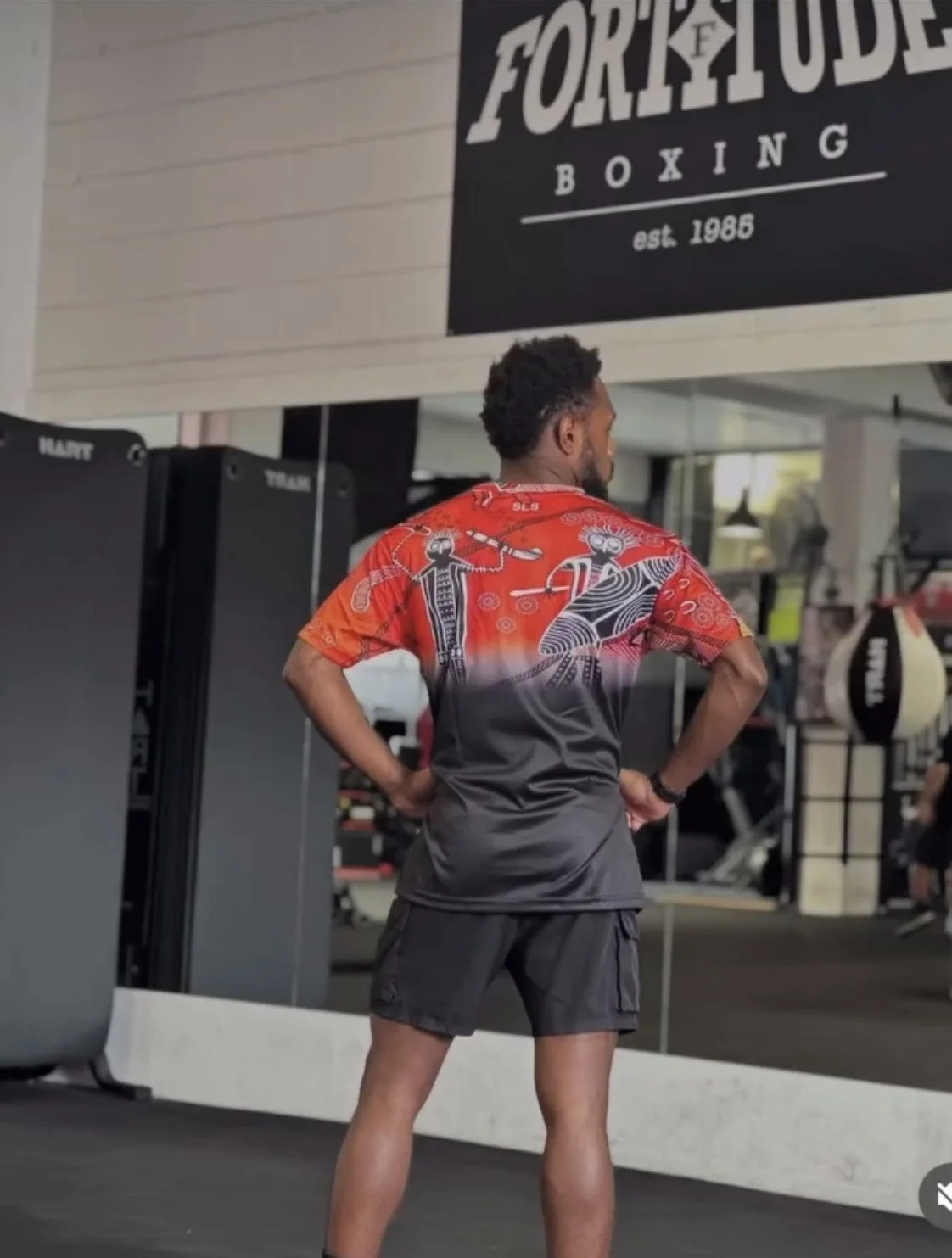 A man standing in a gym wearing black shorts and a colorful shirt with ancient Egyptian figures, facing away from the camera. A sign for Fortitude Boxing, established 1985, hangs above him.