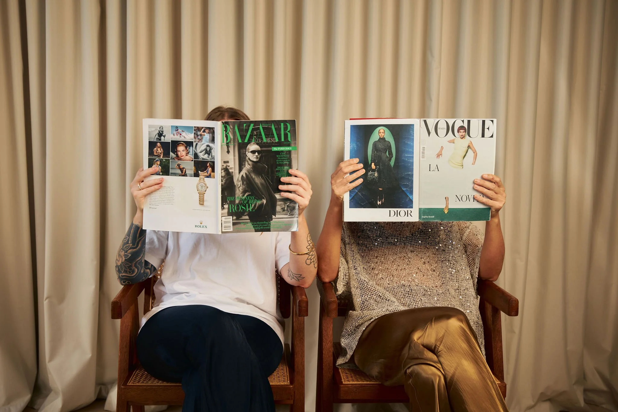 Two women seated on chairs, one reading Vogue and one reading Bazaar