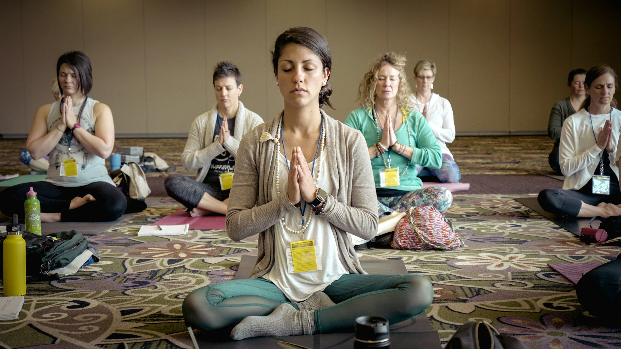 A group of women sitting cross-legged on yoga mats, meditating in a room with neutral-beige walls and floor. They are in a peaceful, focused state with hands in prayer position and eyes closed.