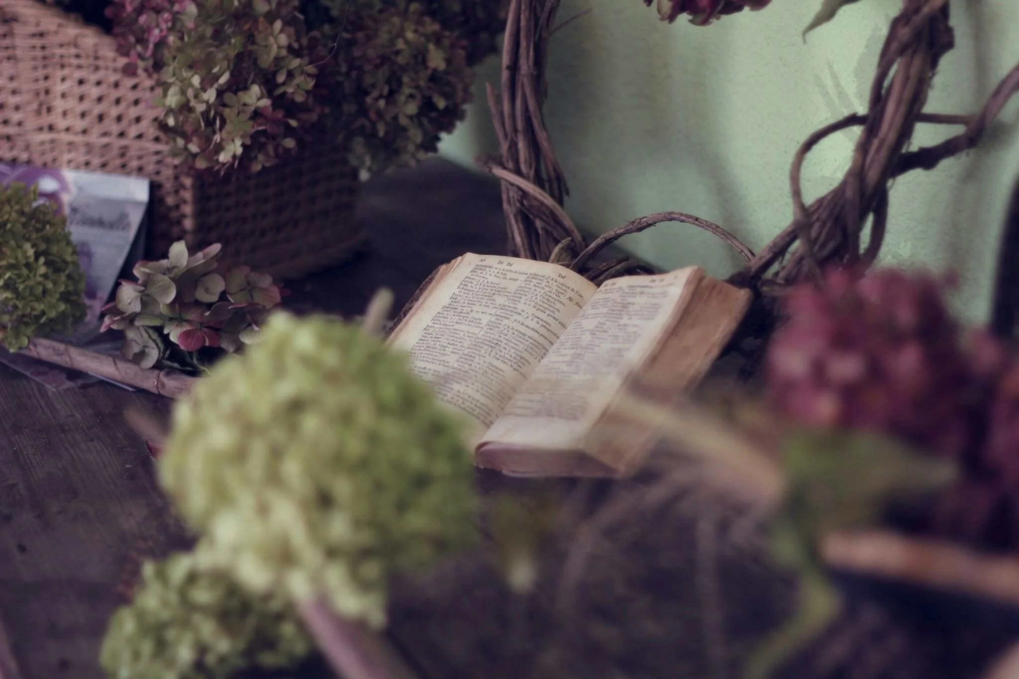 An open Bible placed in a wicker basket surrounded by dried flowers and purple hydrangeas on a dark wooden surface.