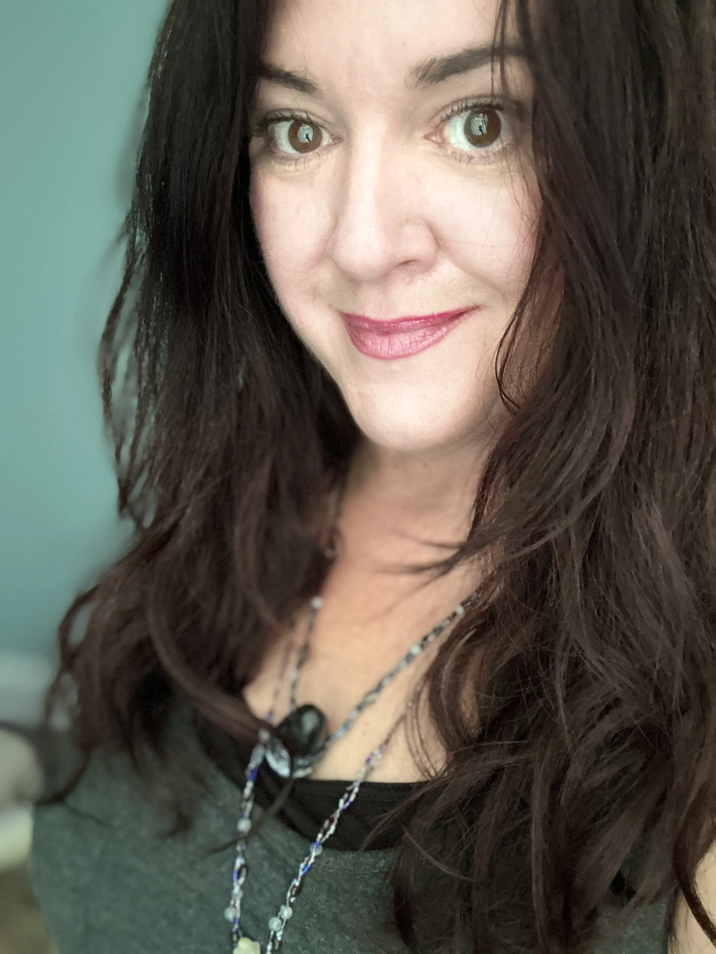 Close-up of a woman with long, wavy dark brown hair, wearing a dark green top and a multi-strand necklace with beads, with a light green background.