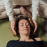 A woman receives a hands-off reiki session from an energy healing practiioner while lying on a green massage table, with her eyes closed and appearing relaxed.