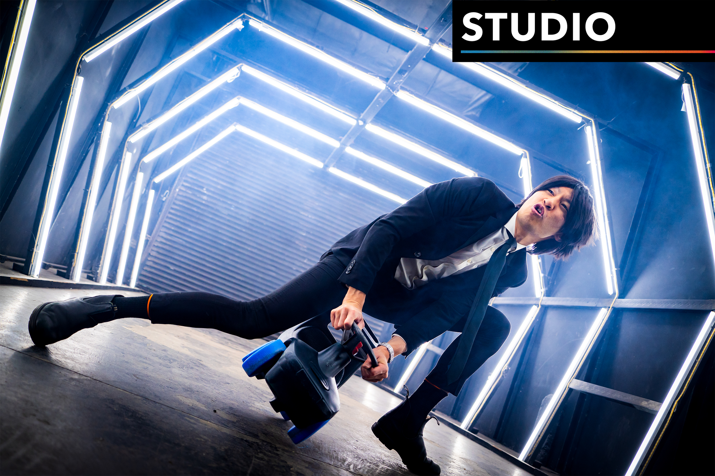 Person in a black suit crouching in a neon-lit studio, holding a prop car. 