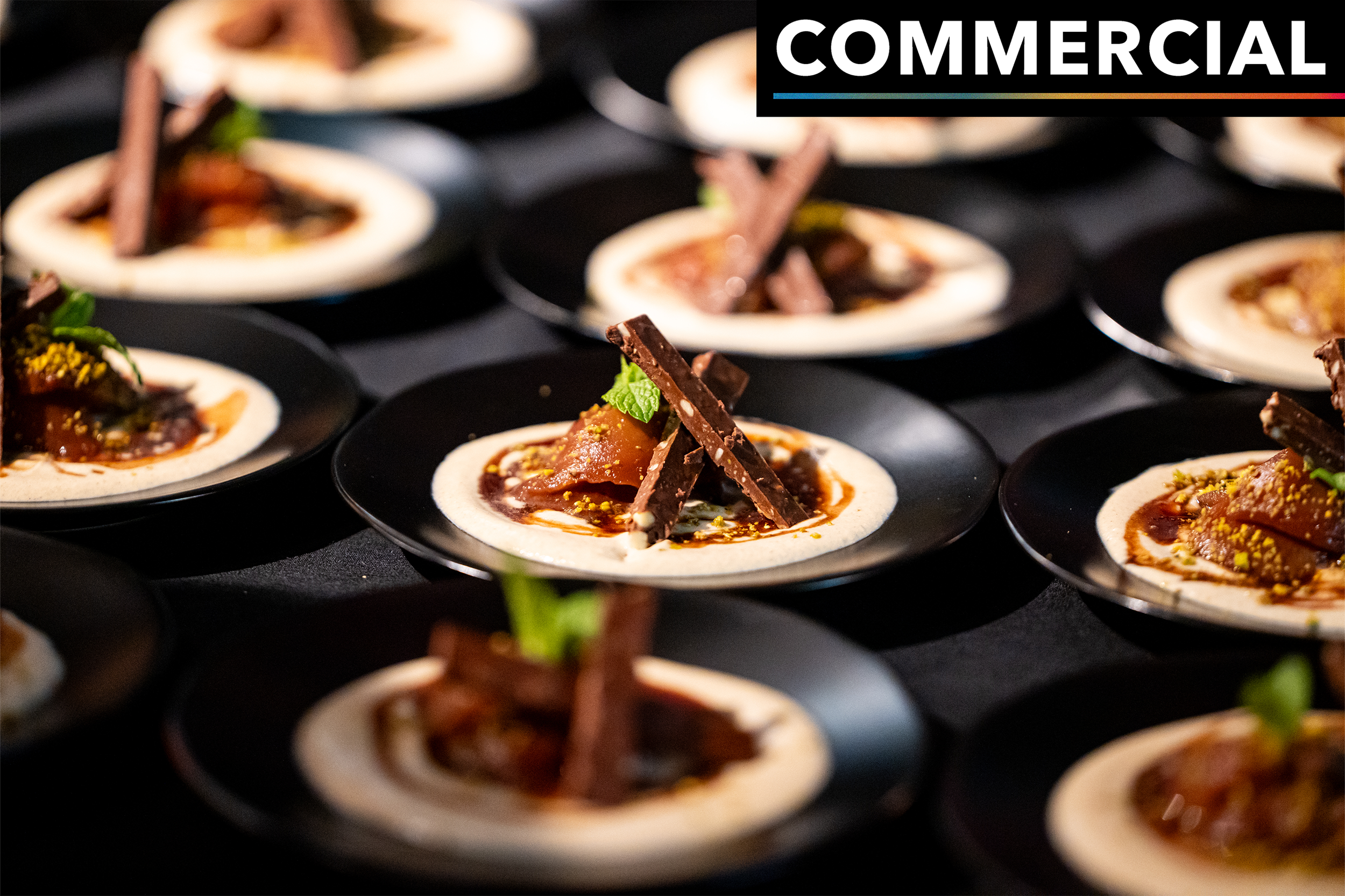 Multiple small black plates each with a dessert consisting of a cookie-like base topped with caramel, chocolate shavings, chopped nuts, and small mint leaves, arranged in a grid pattern.