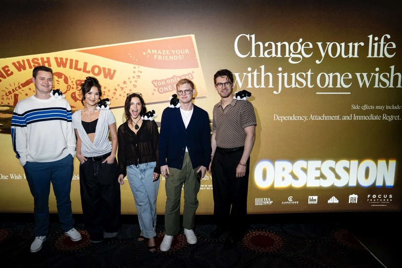 Five young people standing in front of a large movie poster at a promotional event. The poster promotes a film titled 'Obsession' and features the tagline 'Change your life with just one wish.' The group is posing with small plush black and white cats on their shoulders and heads.