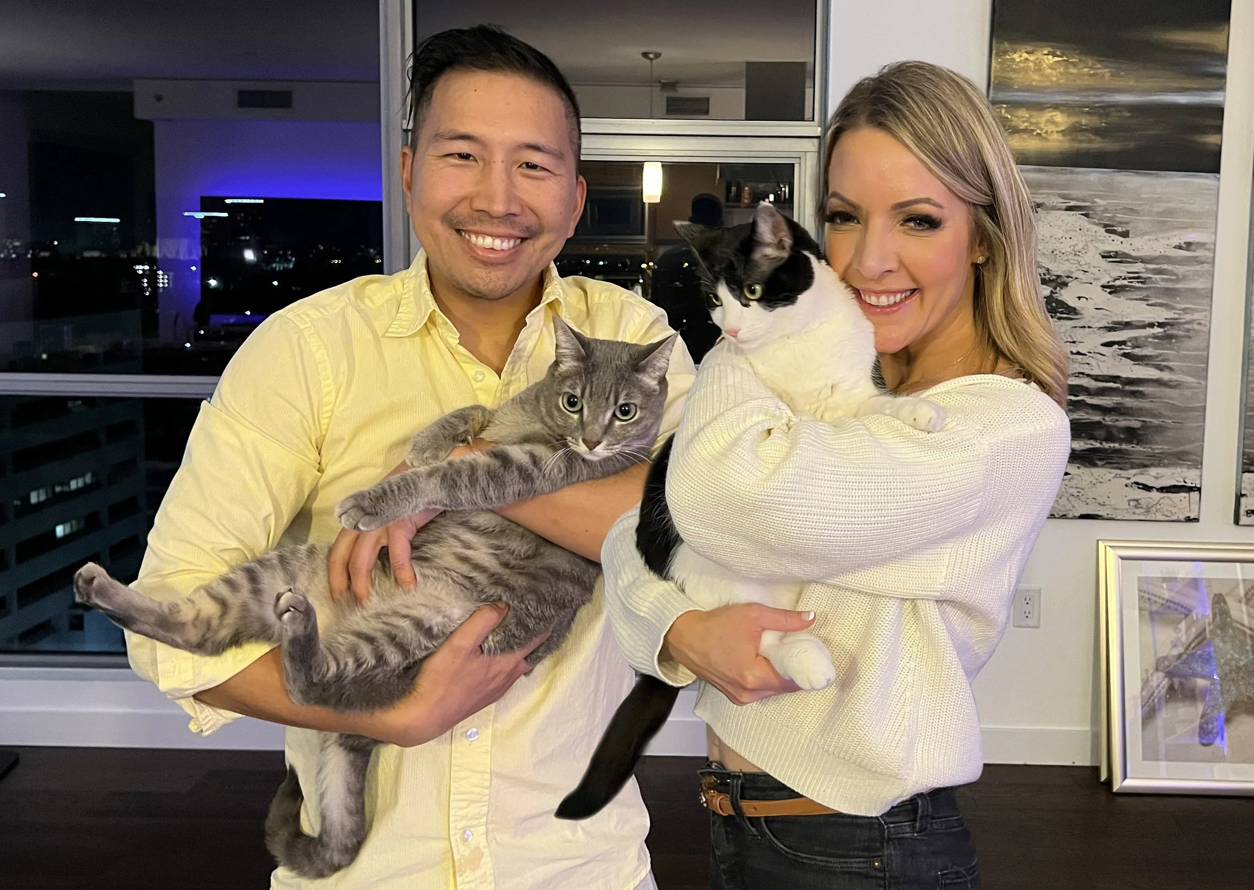 A smiling man in a yellow shirt holds a gray tabby cat, and a smiling woman in a white sweater holds a black and white cat, both in a modern apartment with city views at night.