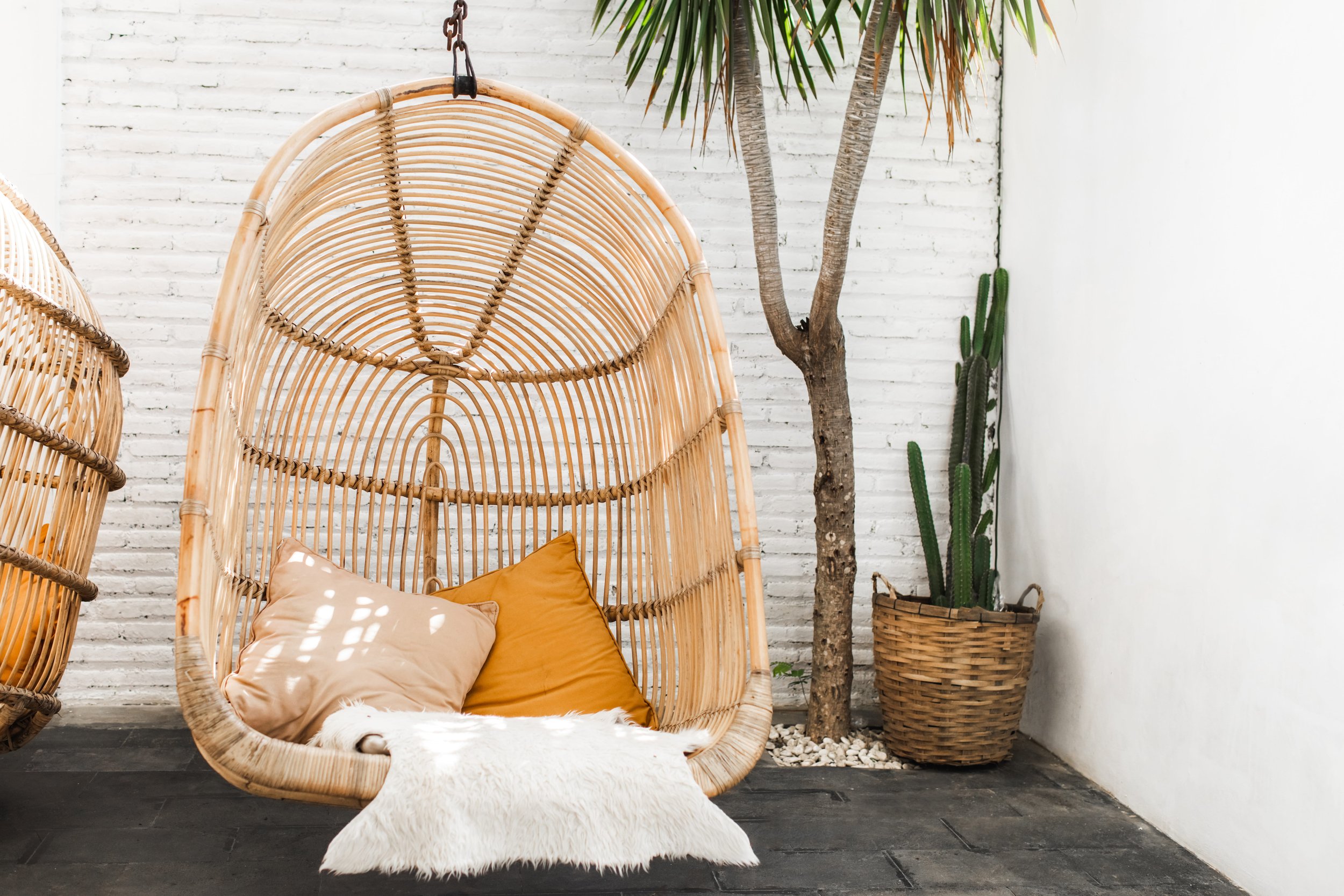 A rattan hanging chair with beige and mustard cushions and a white furry blanket, set against a white brick wall with a potted cactus and a small tree with green leaves.