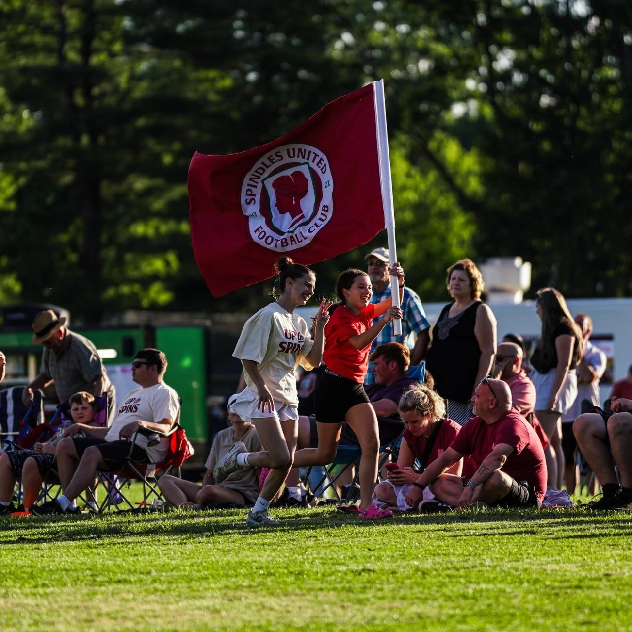 Two people running with a red Spindles United Football Club flag during a sports event, surrounded by spectators on a sunny day.