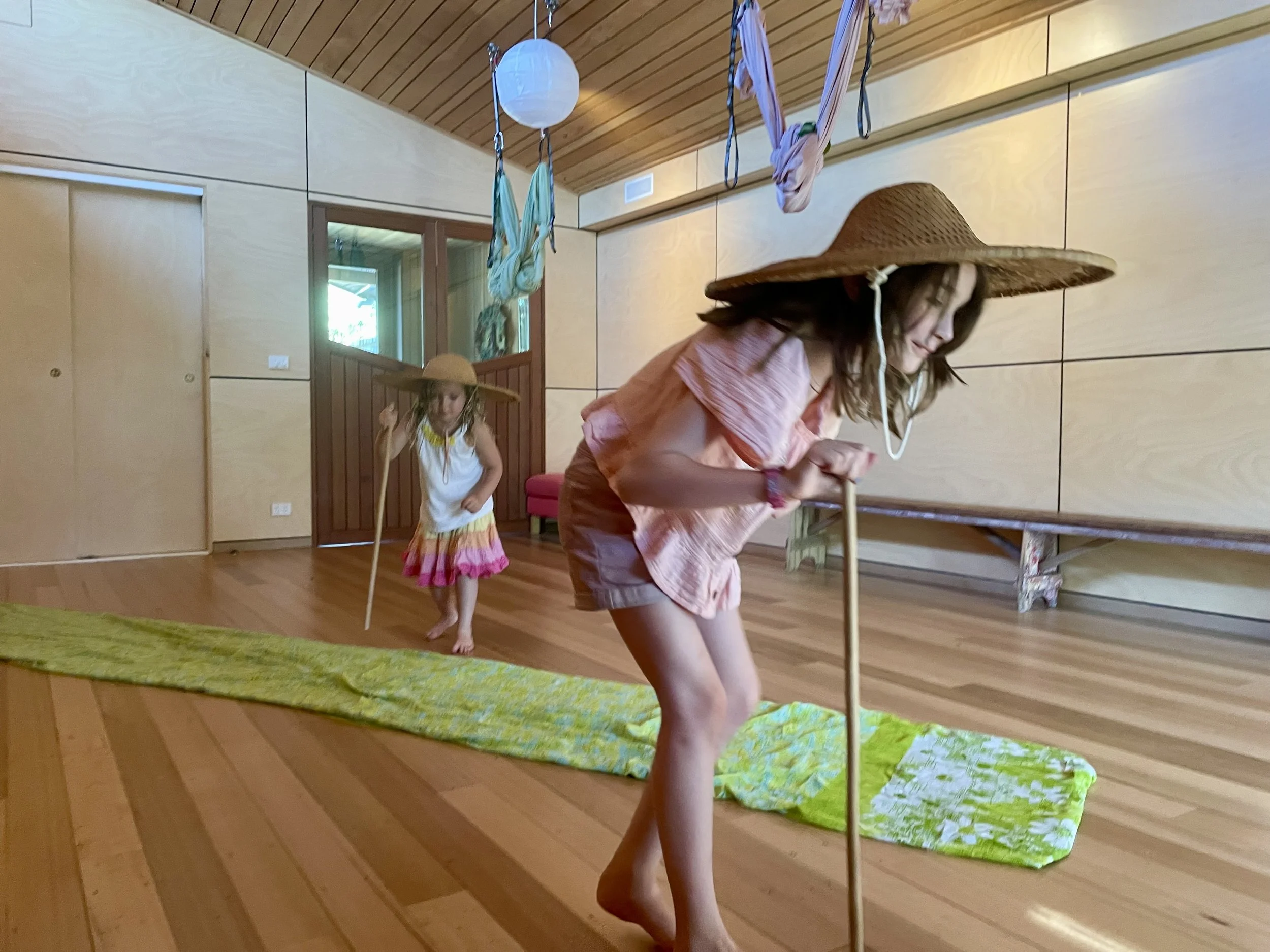 Two young girls wearing wide-brimmed sun hats and summer clothing dancing indoors on a wooden floor with a green mat, cotton candy, and hanging fabric or scarves.