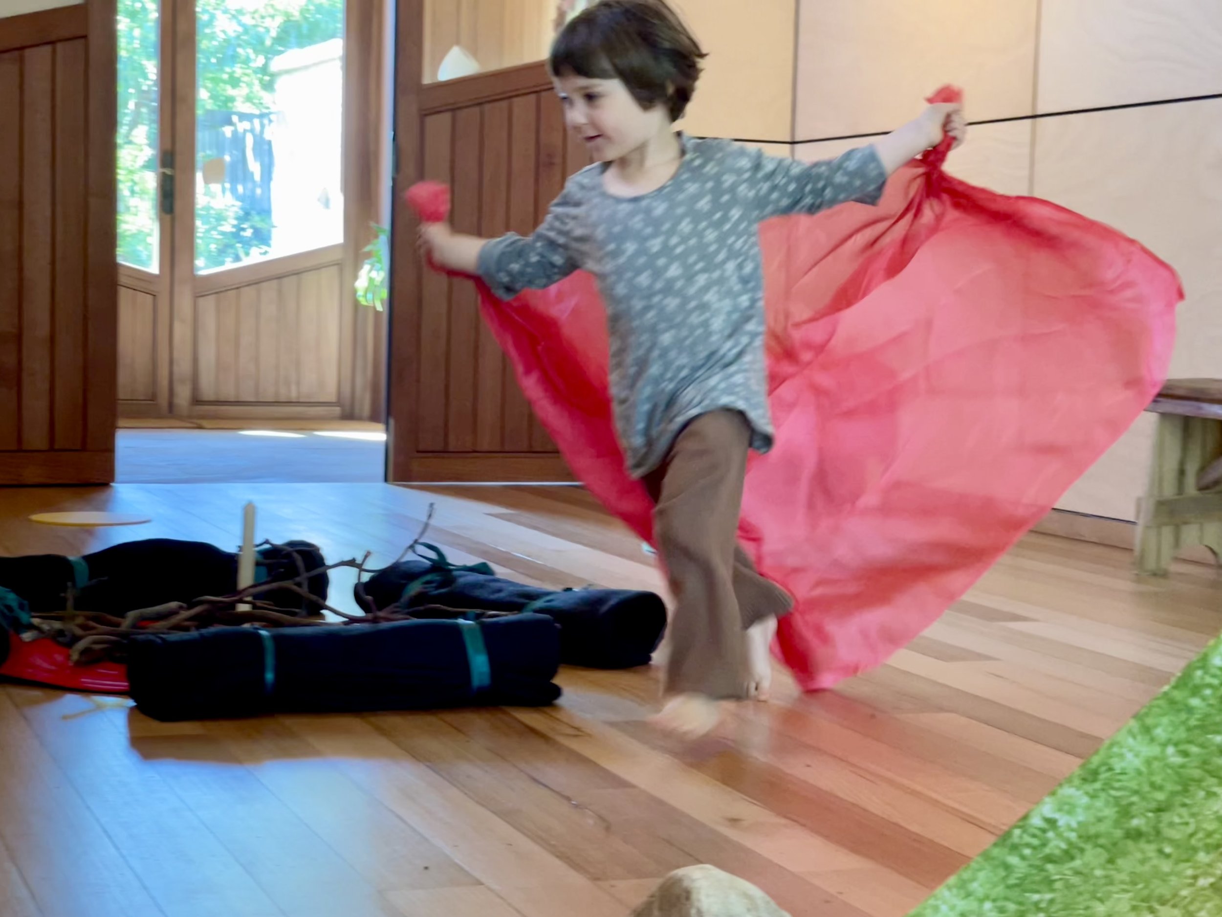 A young child dancing indoors with a red fabric flowing behind them, wearing a gray shirt with white patterns and brown pants, on a wood floor near a doorway.
