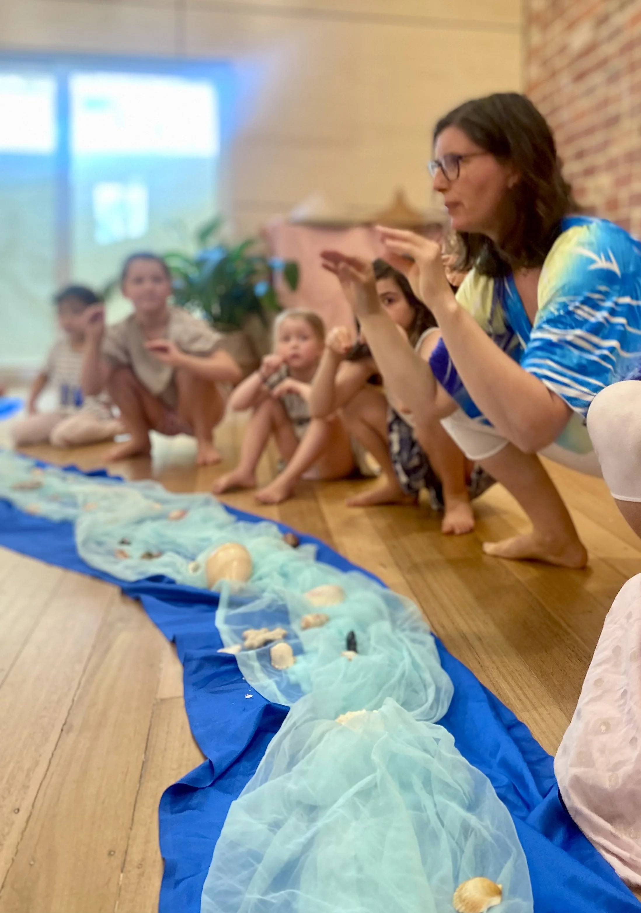 A woman kneeling on the floor surrounded by children, gesturing with her hands as they sit on the floor listening. There is a blue cloth with shells and fabric laid out in front of them, resembling an underwater scene.
