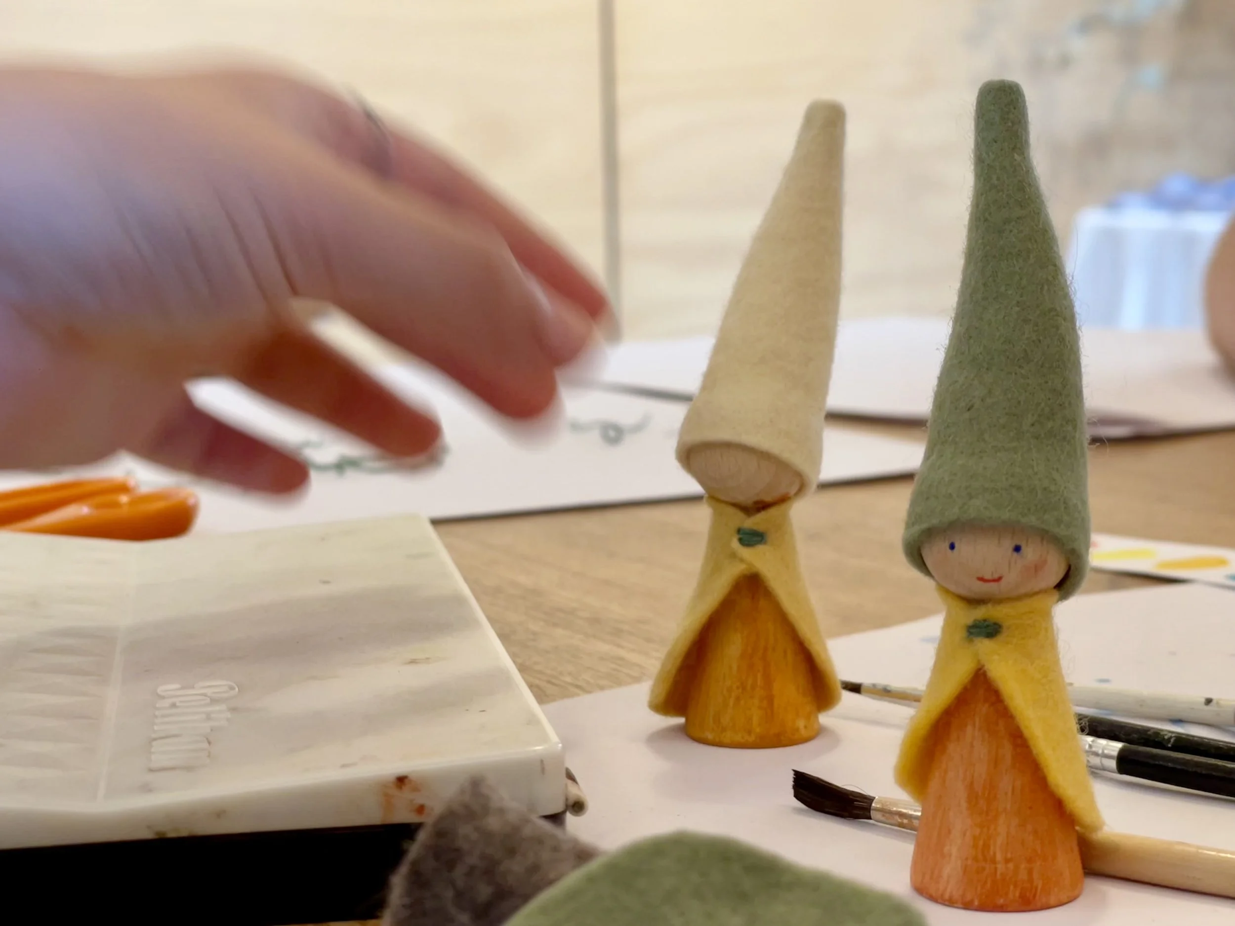 Two small handcrafted dolls with cone-shaped hats, one green and one yellow, standing on a table surrounded by art supplies.