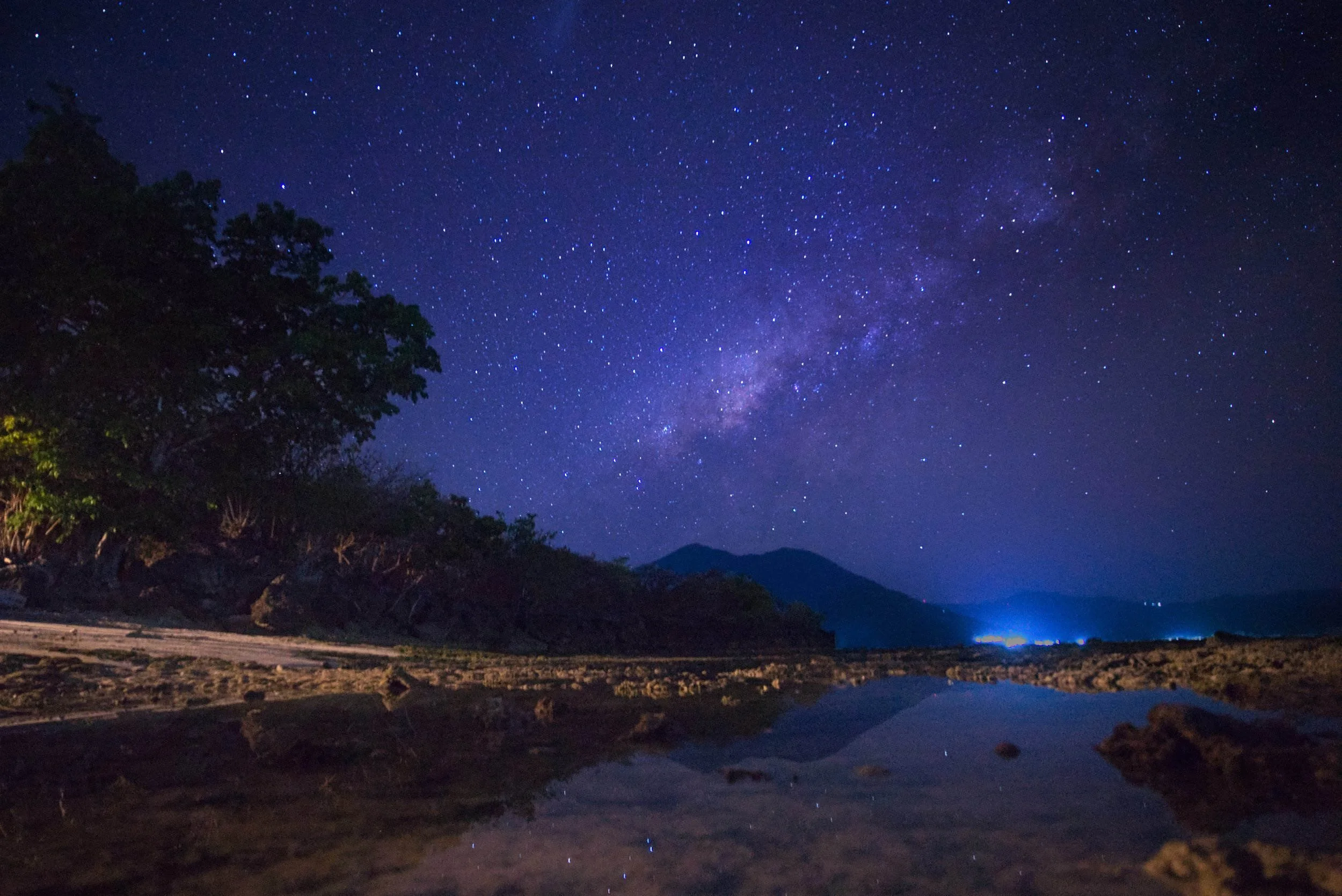 Landscape astrophotography photo of the Milky Way above a volcano and rocky sandy tropical beach at night in the Alor Archipelago, Indonesia.