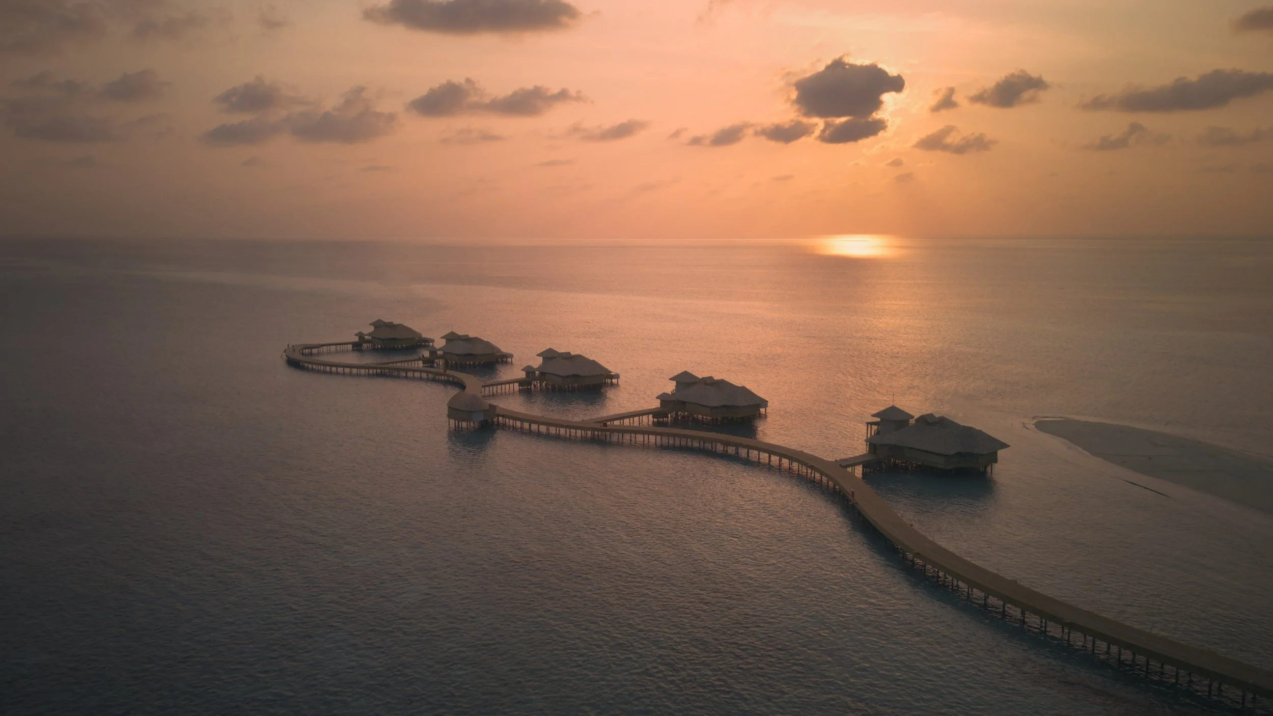 Drone photo of over water villas surrounded by calm ocean at a luxury resort in the Maldives with a beautiful orange sunset.