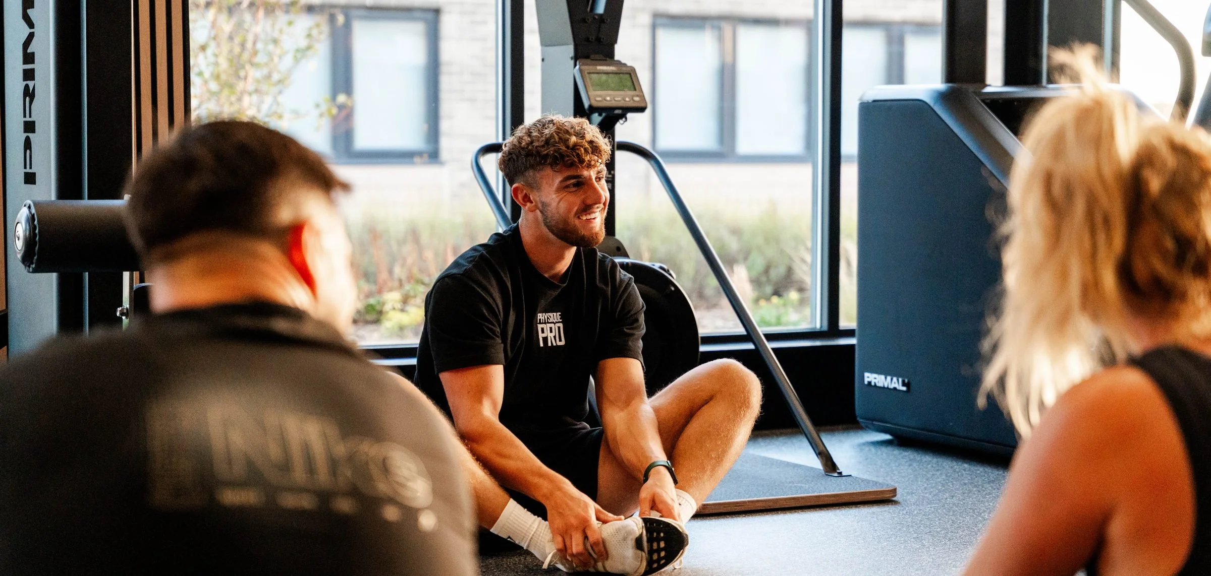 Physique Pro coach stretching on a mat with two members beside the windows at Great Park