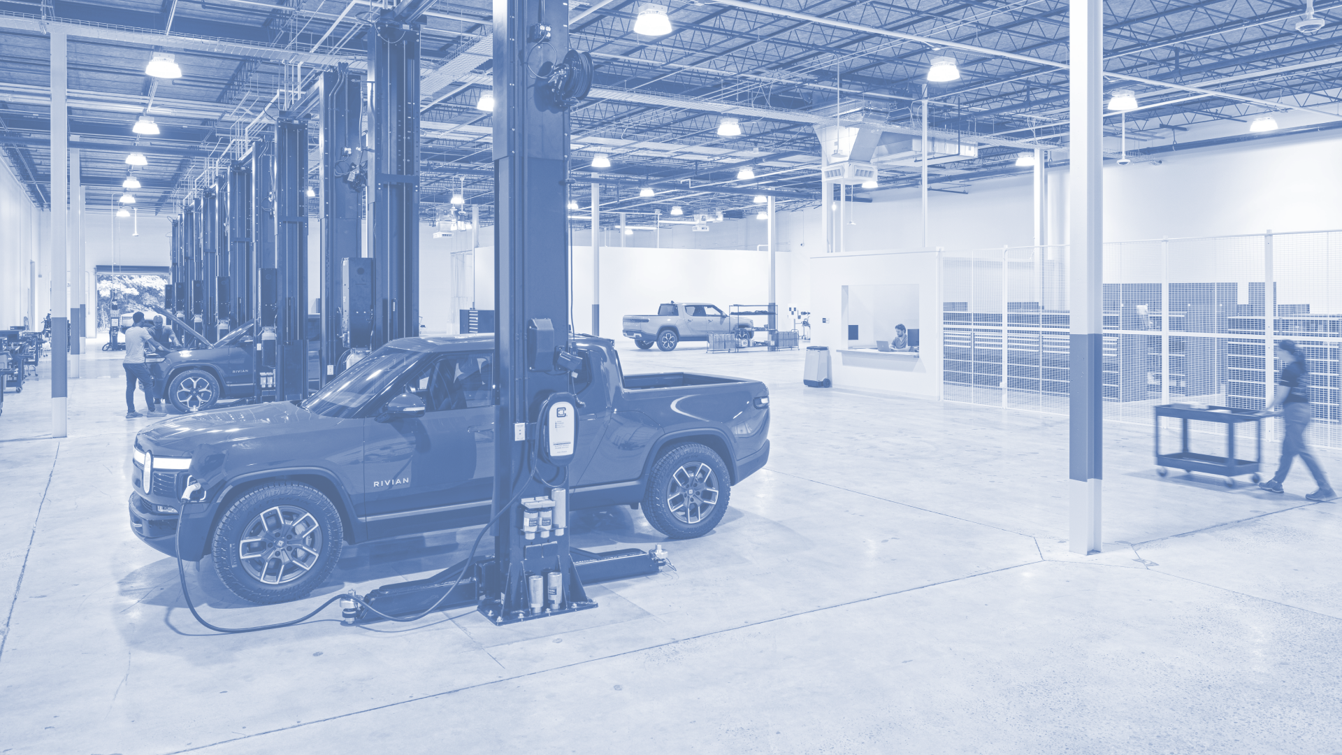 Inside an electric vehicle manufacturing or maintenance facility with several Rivian vehicles and people working.