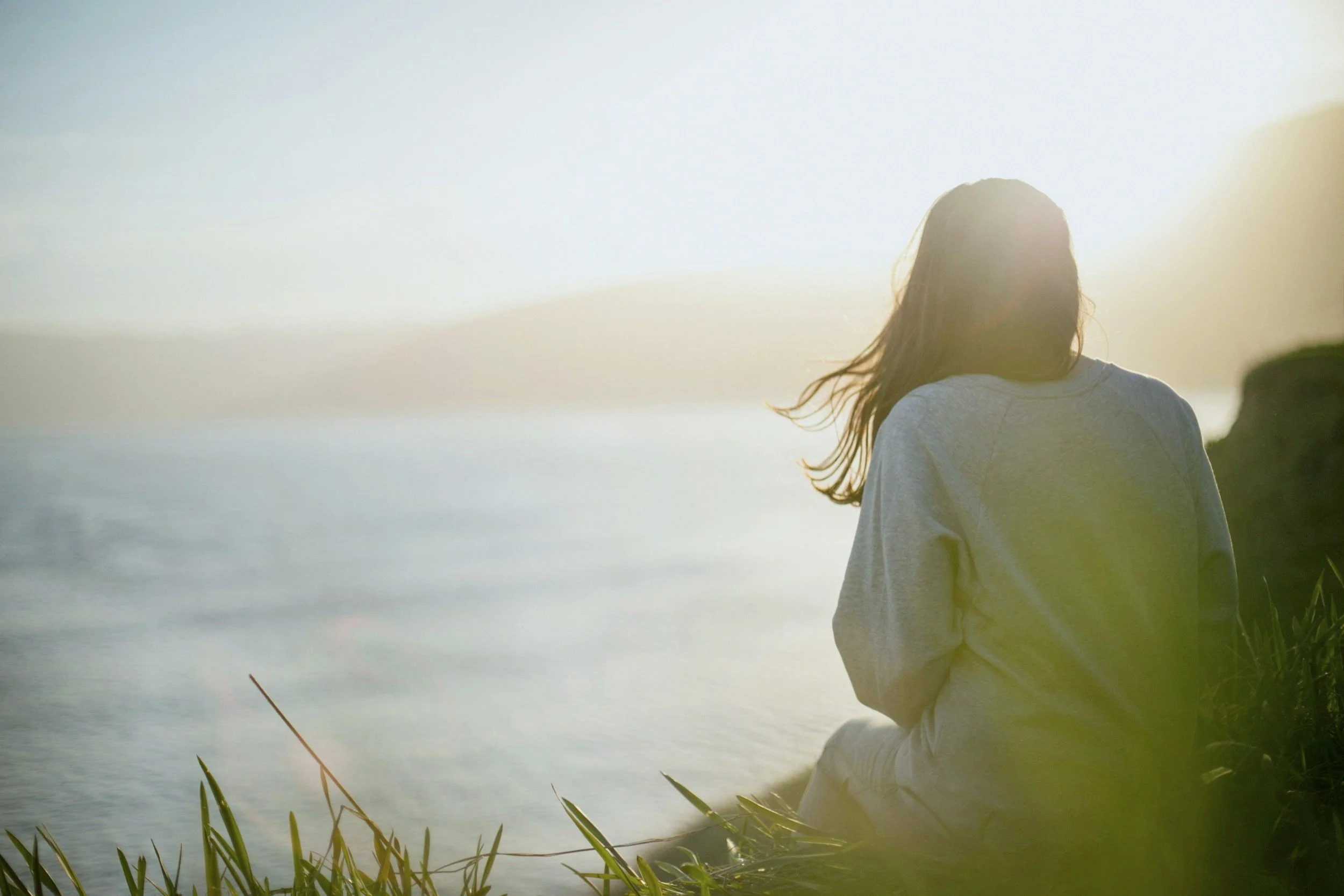 Eine junge Frau steht am Wasser, angelt und blickt in die Ferne, während die Sonne im Hintergrund scheint.