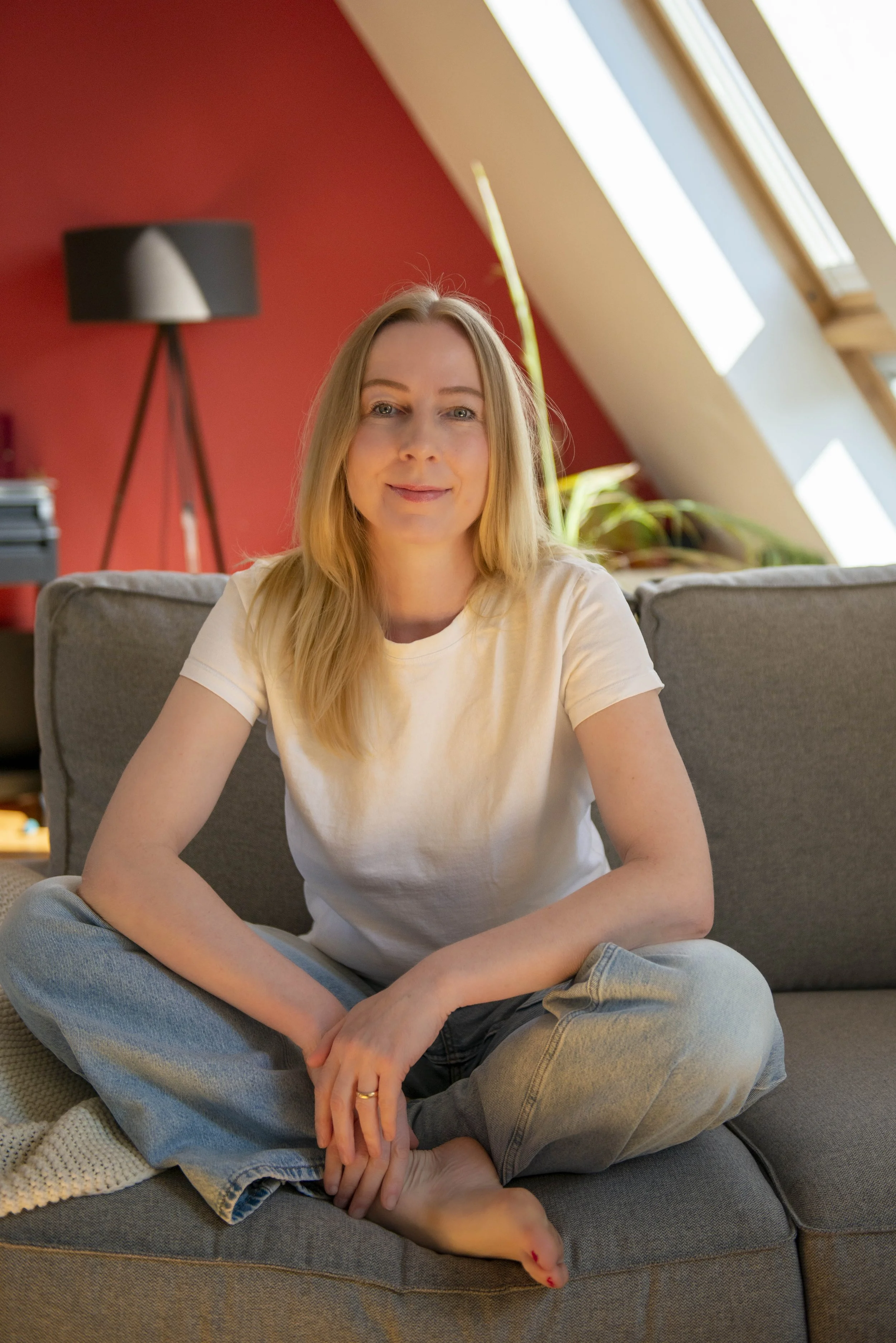 Meike Sieveking sitzt entspannt auf einem grauen Sofa in einem Raum mit roter Wand und Dachfenstern, lächelt in die Kamera.
