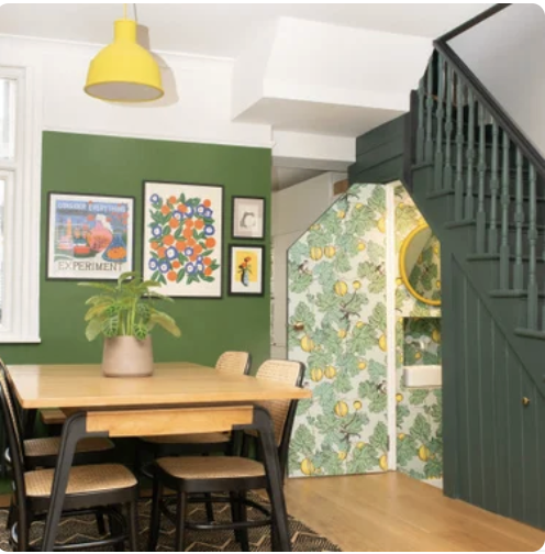Dining area with a green accent wall decorated with framed artwork, a wooden table with chairs, a large potted plant, green staircase with wallpapered wall under the stairs featuring lemons and leaves, and a yellow pendant light.