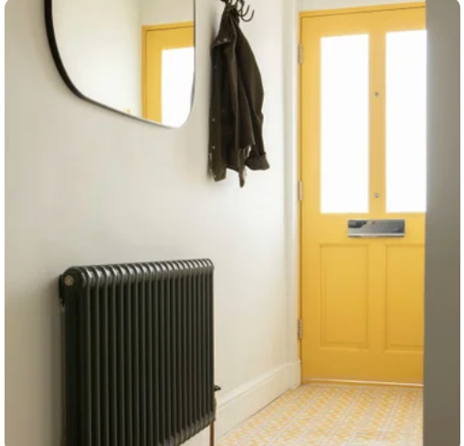 Entryway with a yellow door, black coat hanging on a hook, radiator on a white wall, and a mirror.