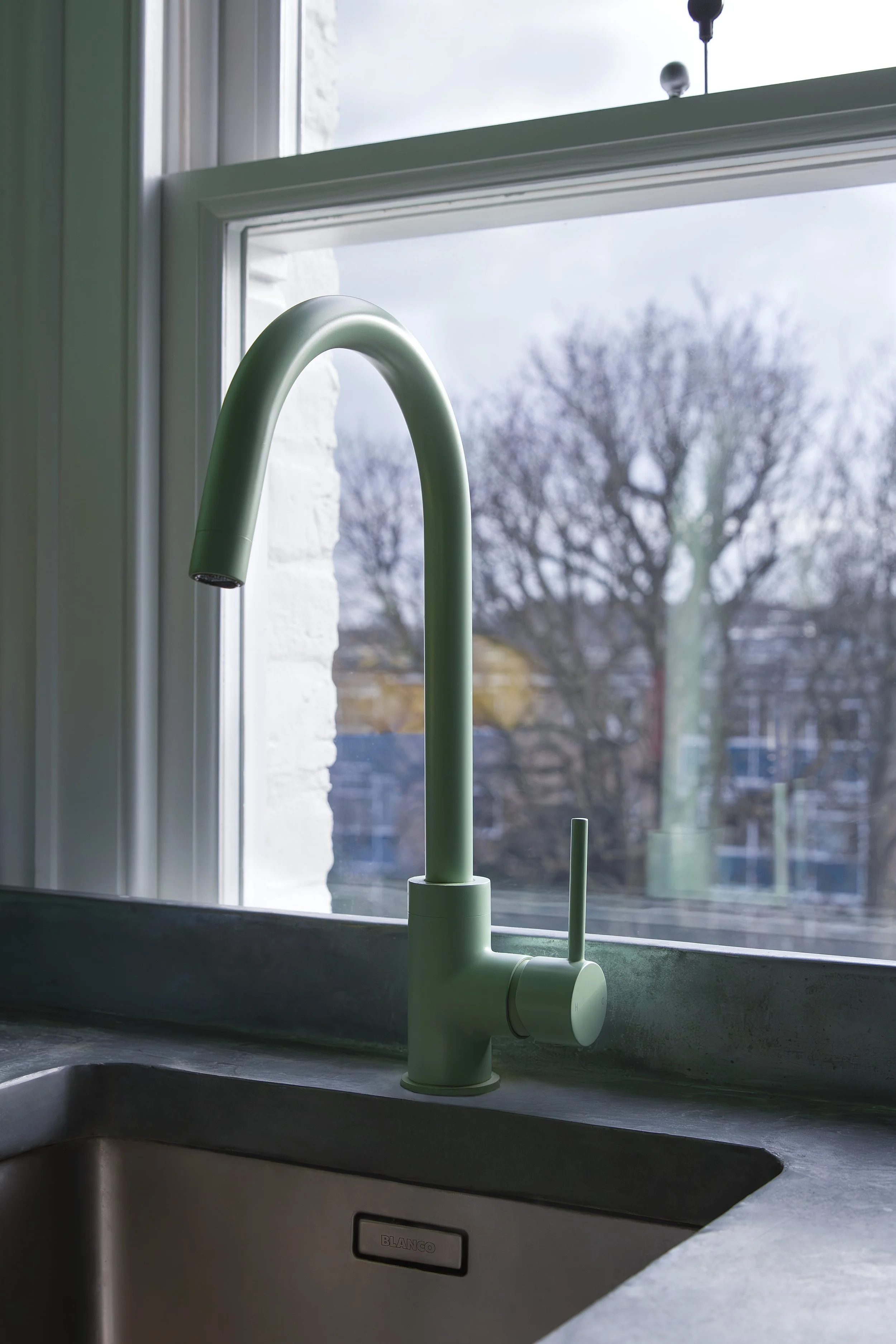 Close-up of a modern green kitchen faucet mounted on a gray countertop next to a stainless steel sink, with a view of a cloudy outdoor scene through a large window.
