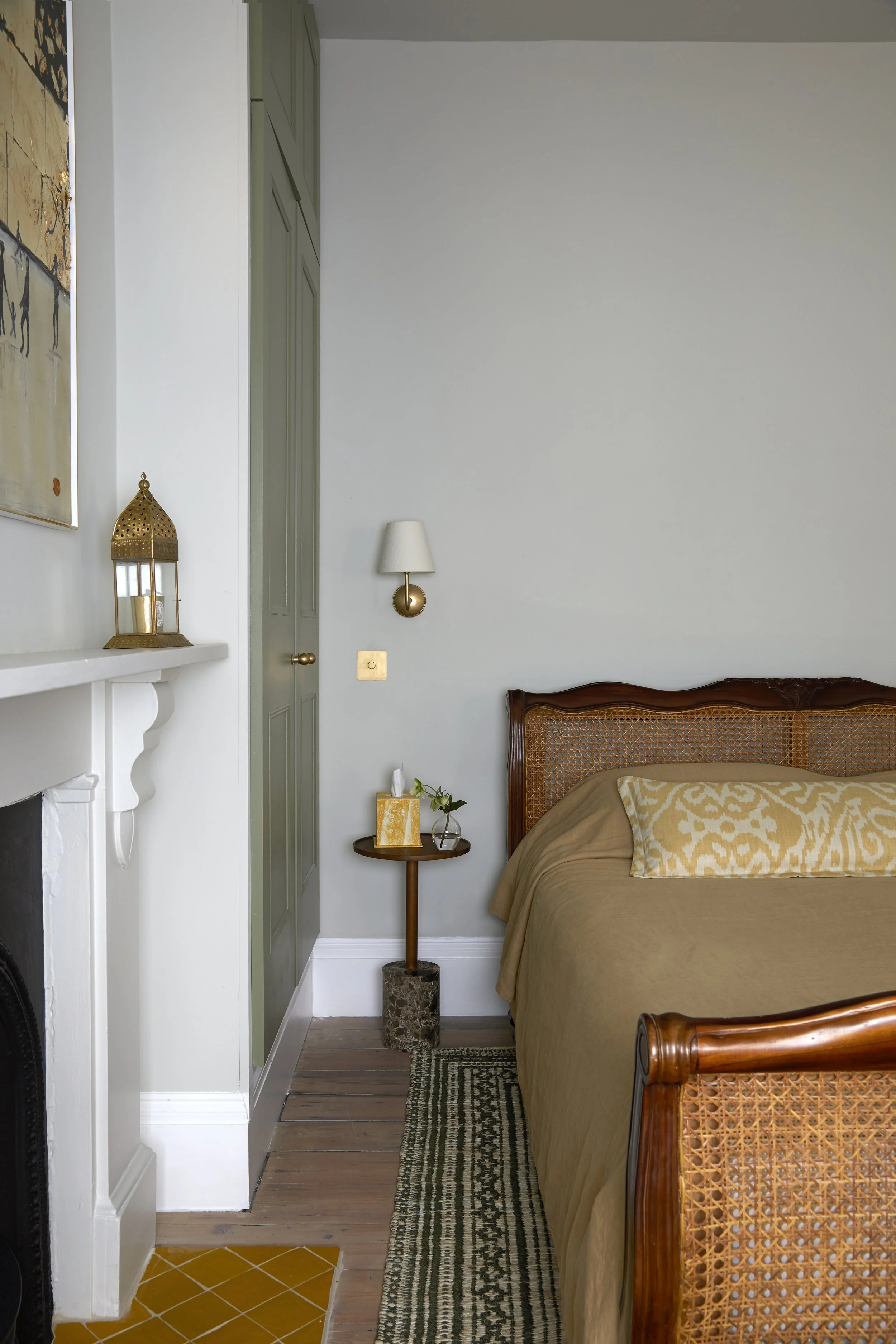 A bedroom corner with a wooden bed, bedside table with flowers, tissue box, wall-mounted light, and a green built-in closet door. Part of a patterned rug, fireplace with decorative items, and artwork are visible.