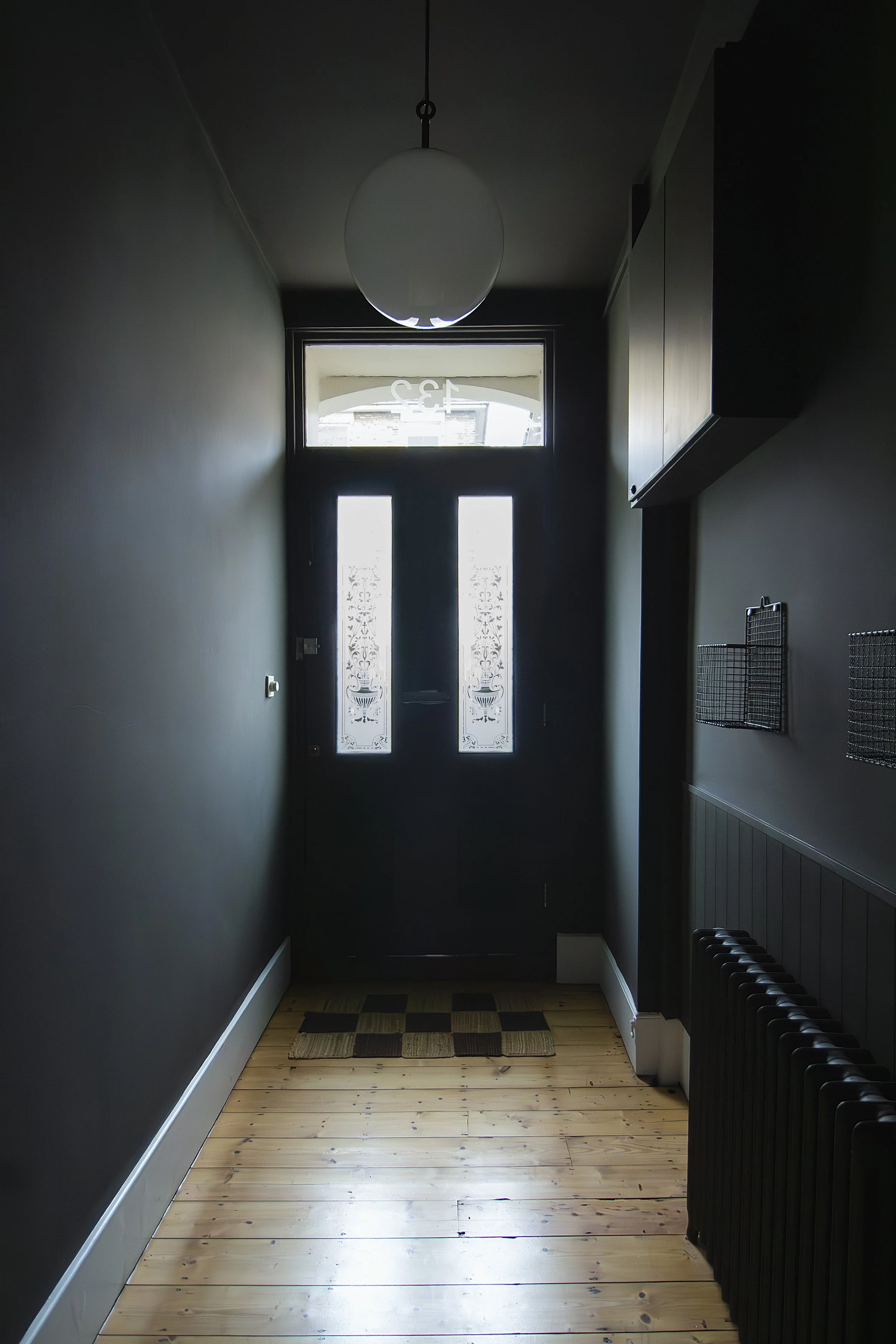 Narrow entryway with dark door featuring decorative glass panels, light wooden floor, dark green walls, small wall-mounted baskets, radiator, and a modern round ceiling light.
