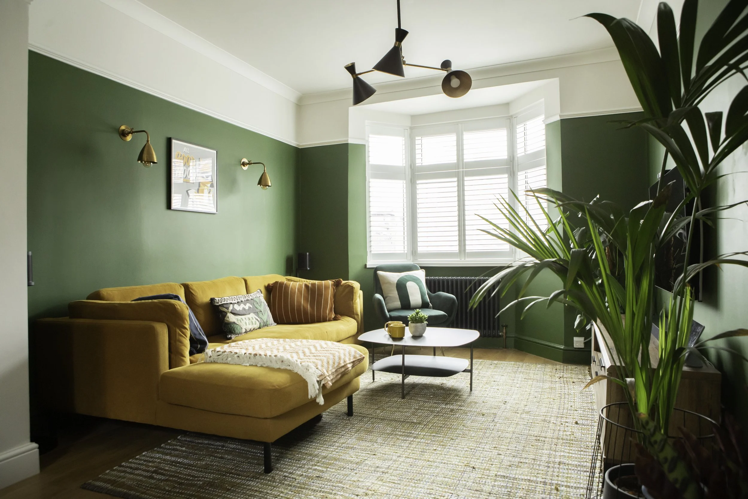 Living room with green walls, yellow sectional sofa with multi-colored pillows, white coffee table, green armchair, large window with white shutters, potted plants, ceiling light fixture, and framed artwork on the wall.