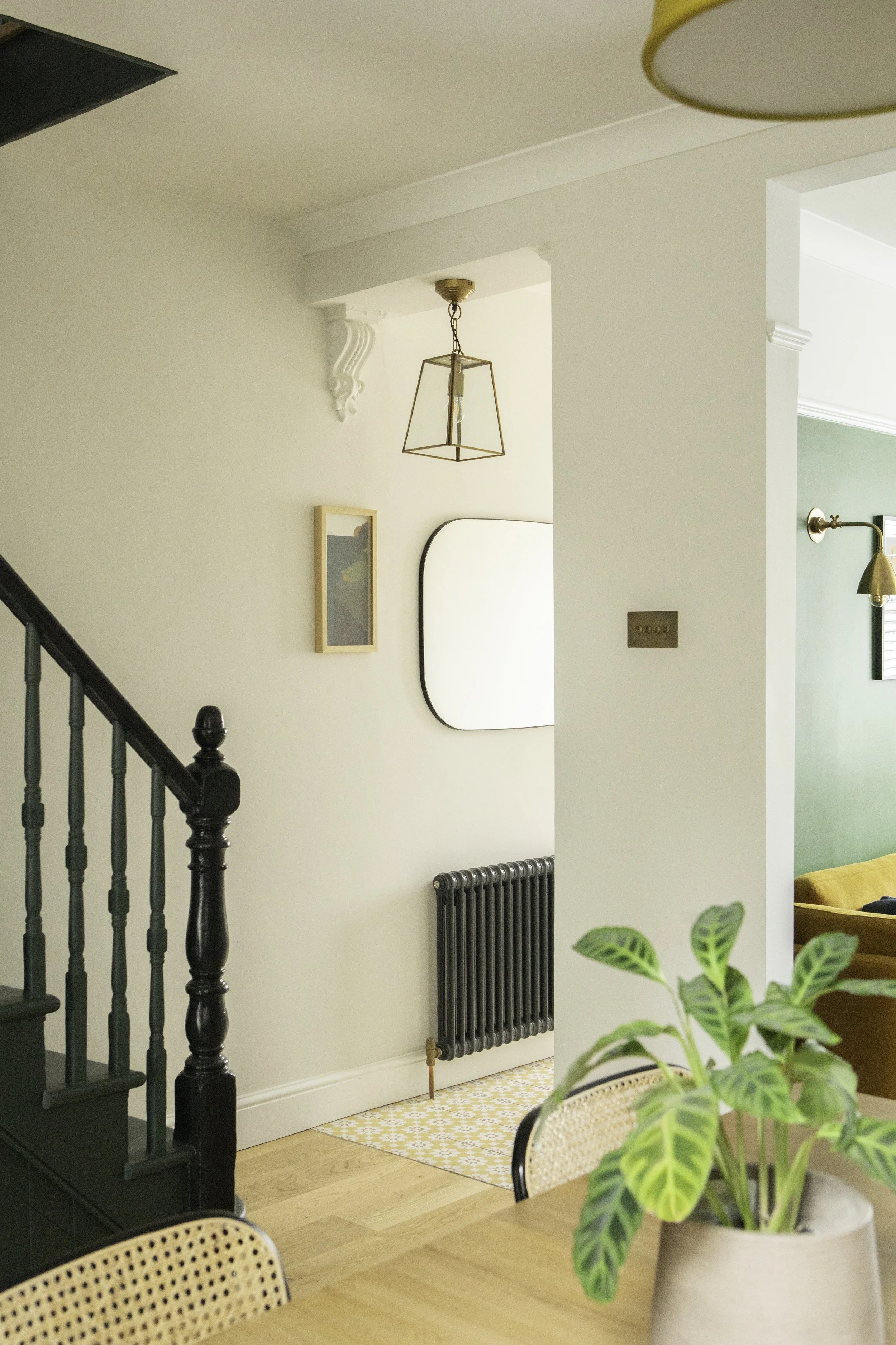 Interior view of a home with a wooden dining table, green houseplant in a beige pot, a staircase with black balusters, a radiator, framed artwork, and a mirror on the wall with a hanging geometric light fixture.