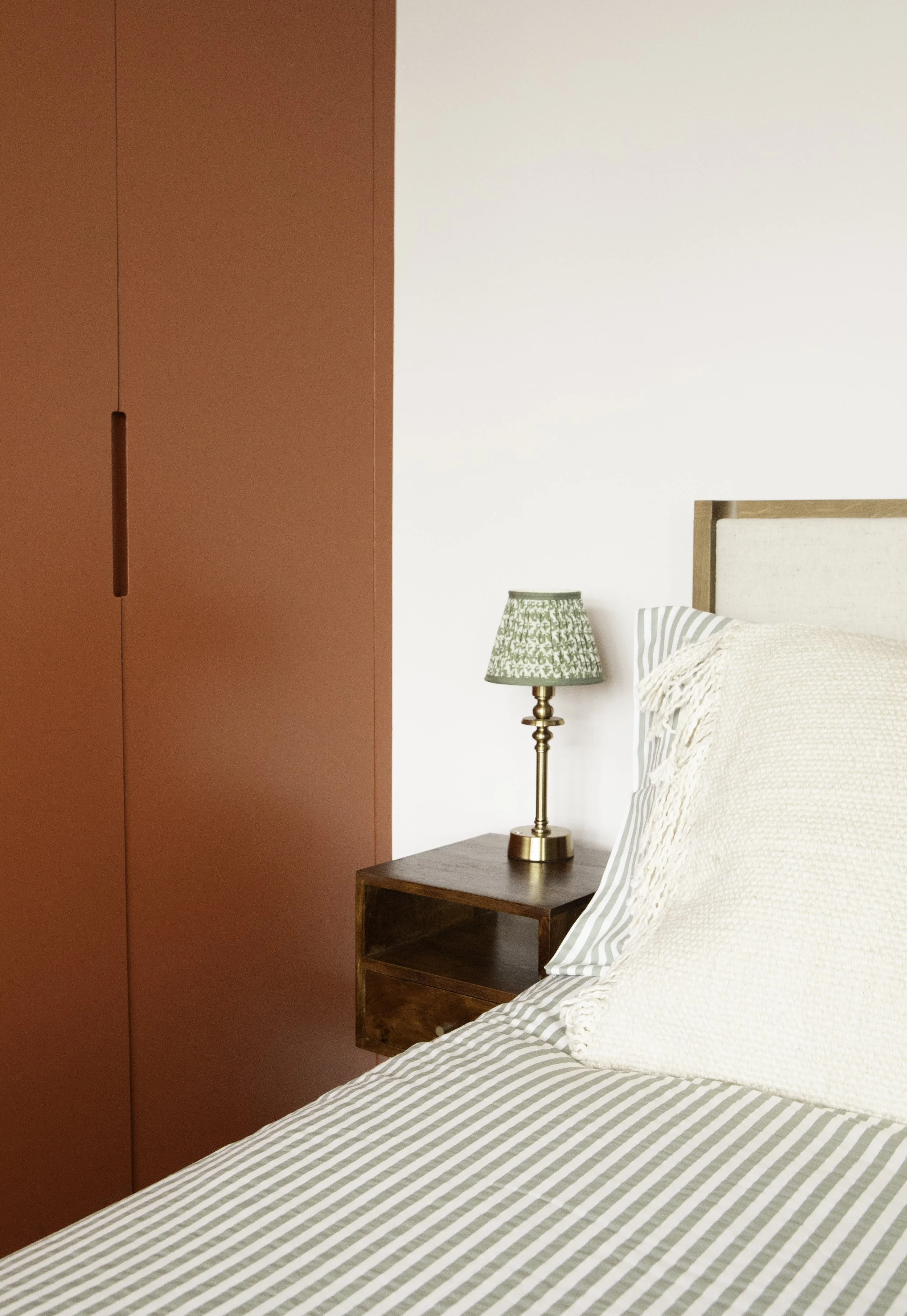 A bedside table with a brass lamp and green patterned shade next to a bed with striped sheets and a cream pillow, against a white wall with a large  wardrobe painted in Farrow and Ball
